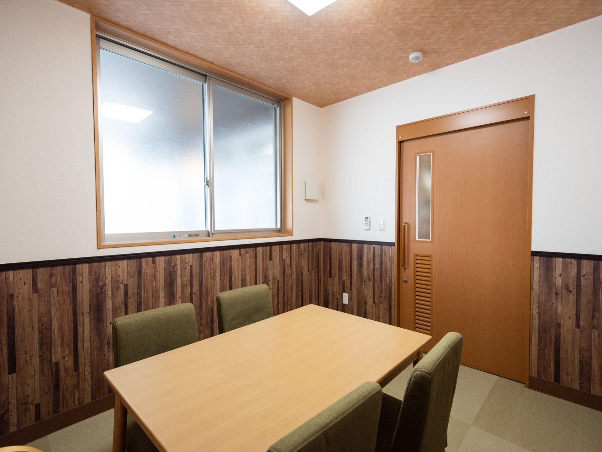 部屋は窓が１つあり、壁面の上部が白色で下部が木目調の個室である。部屋の中央には、四角いテーブルに椅子が2脚ずつ向かい合って配置されている。