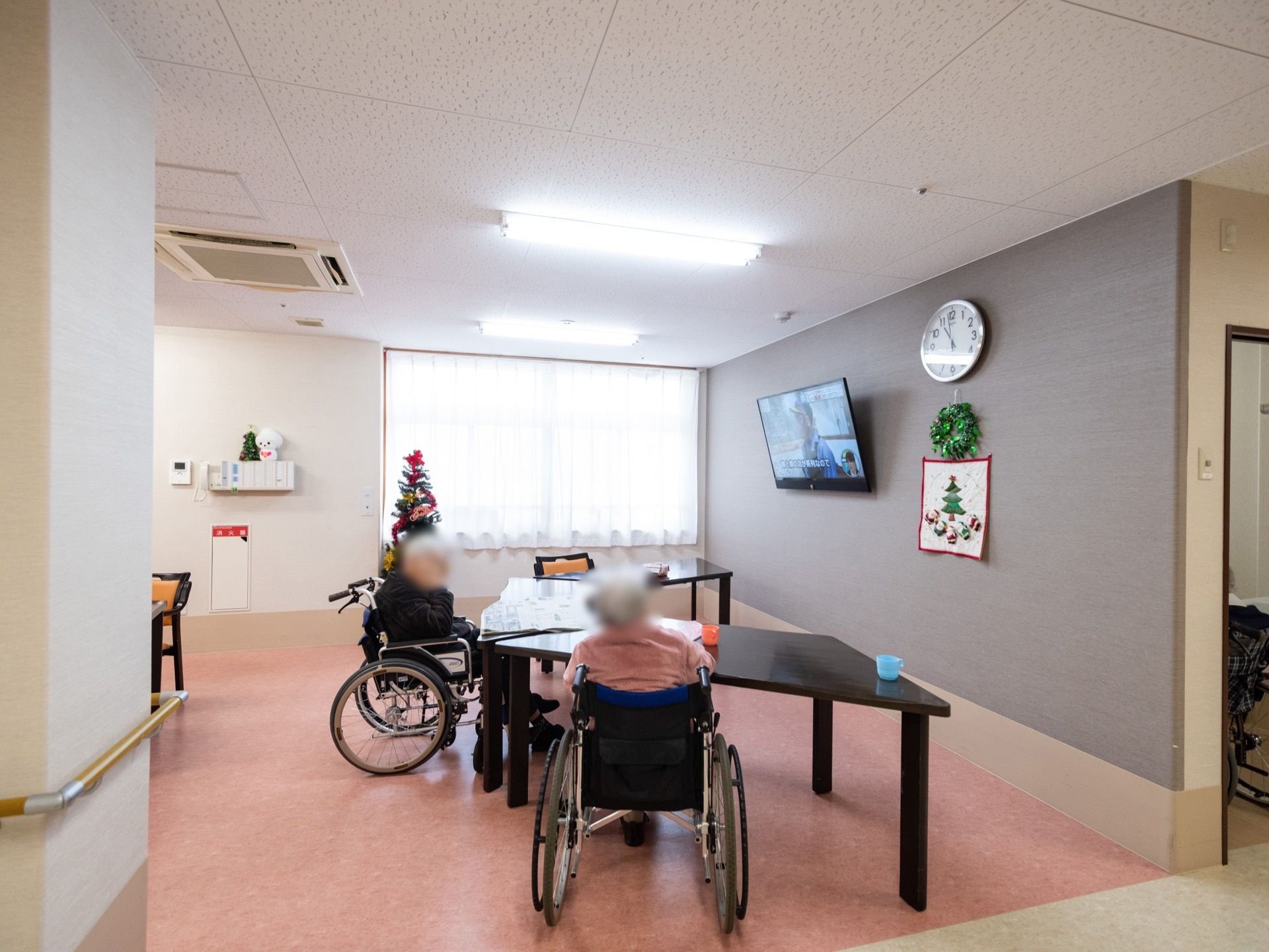 車椅子をご利用の入居者様が２人、同じテーブルに着席されている。向かい側の壁面にはテレビが設置され、横に時計と手作り作品が飾られている。