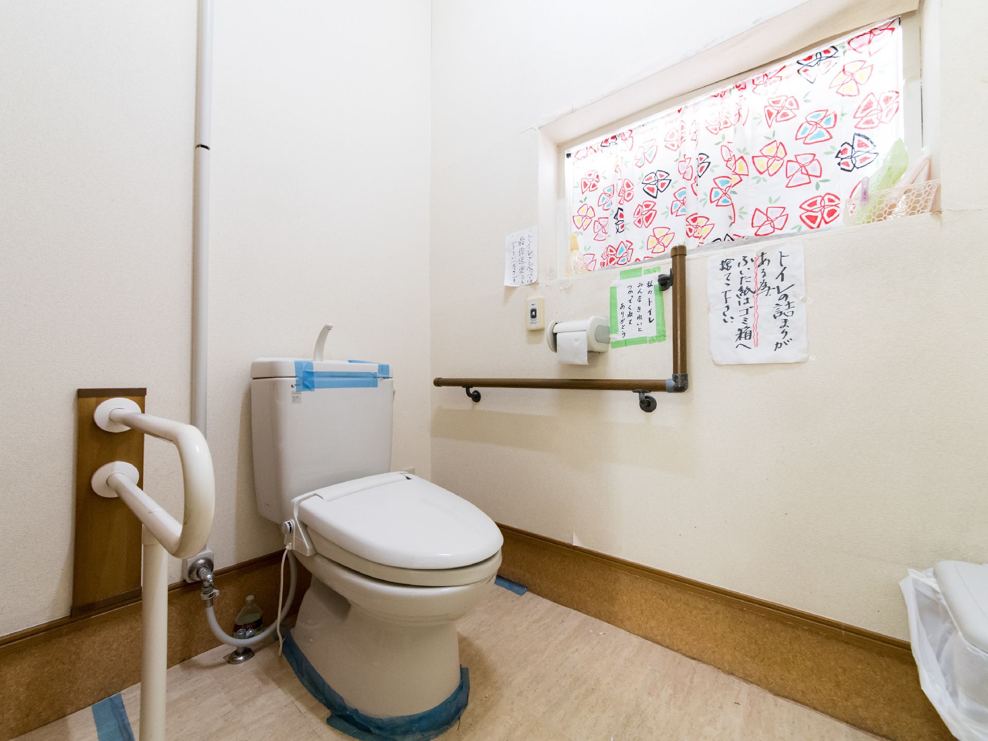 トイレには、利用者の立ち座りの動作を補助するL字型と可動式の手すりが備え付けられている。注意書きが壁に貼られている。