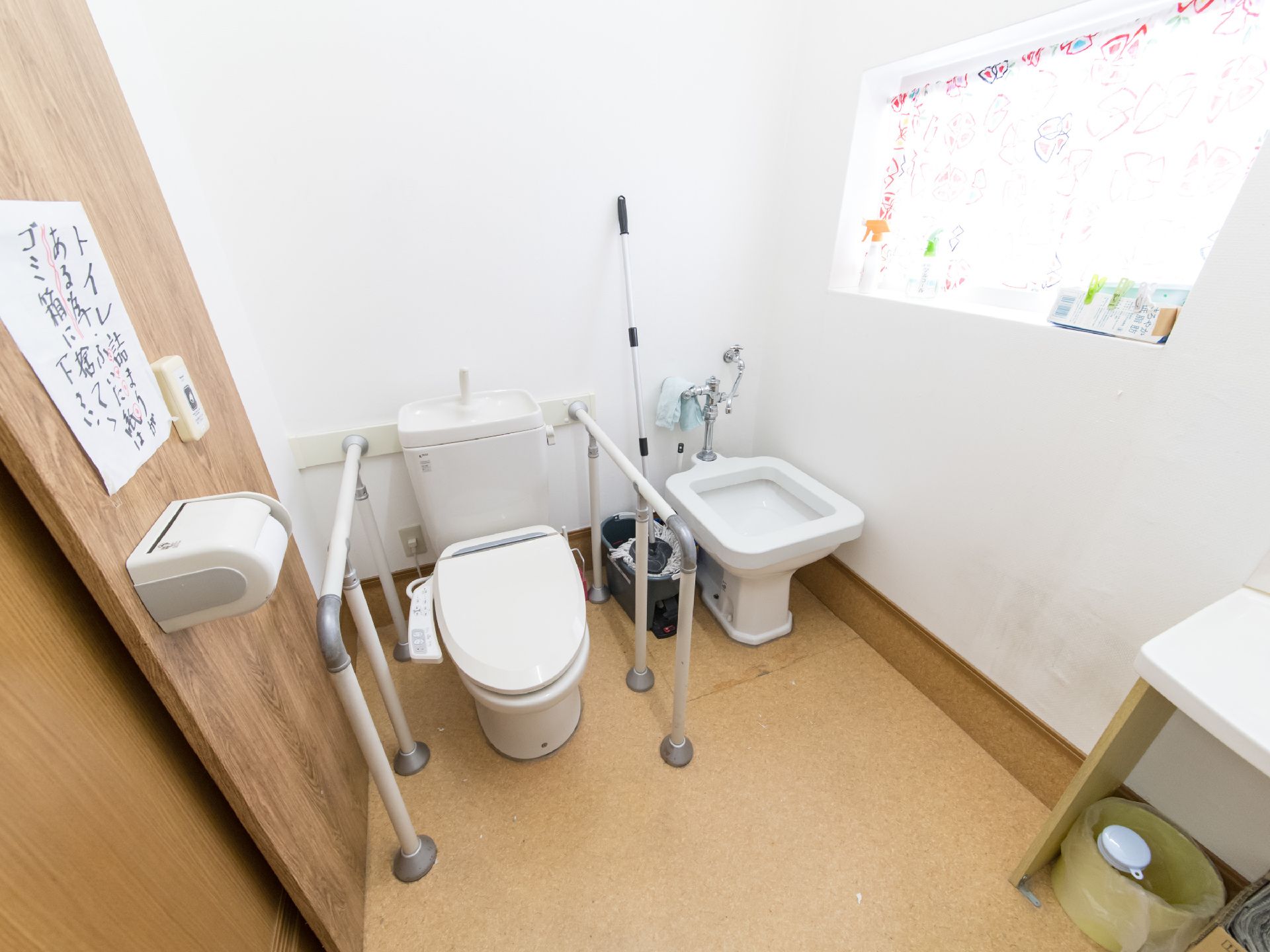 洋式便器の隣にビデが設置されているトイレである。両手で掴まれるよう手すりが取り付けられている。掃除用具が置かれている。