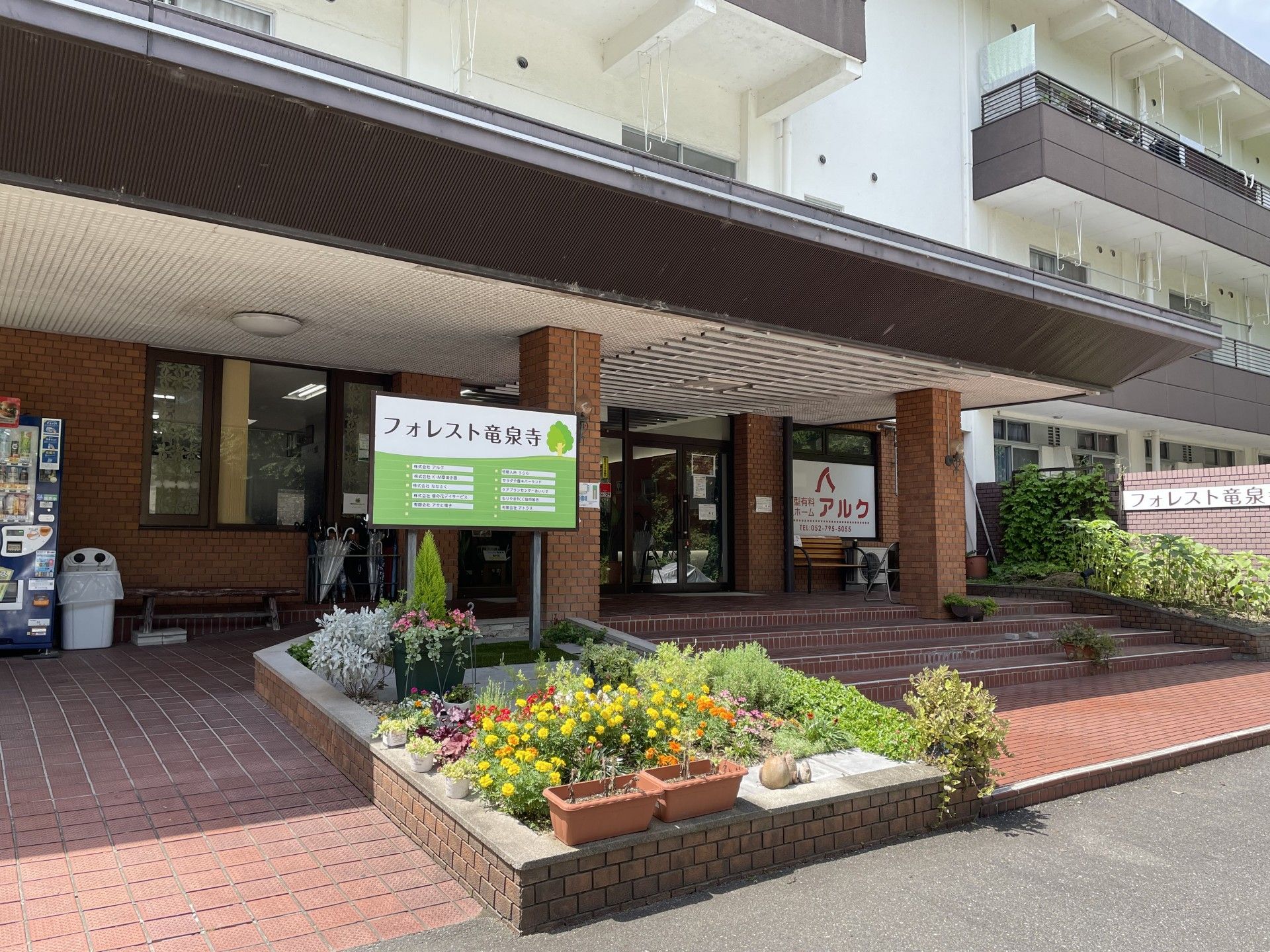 舗装された敷地に、３階建ての大きな建物がある。建物の周りには植栽があり、前は駐車スペースになっている。