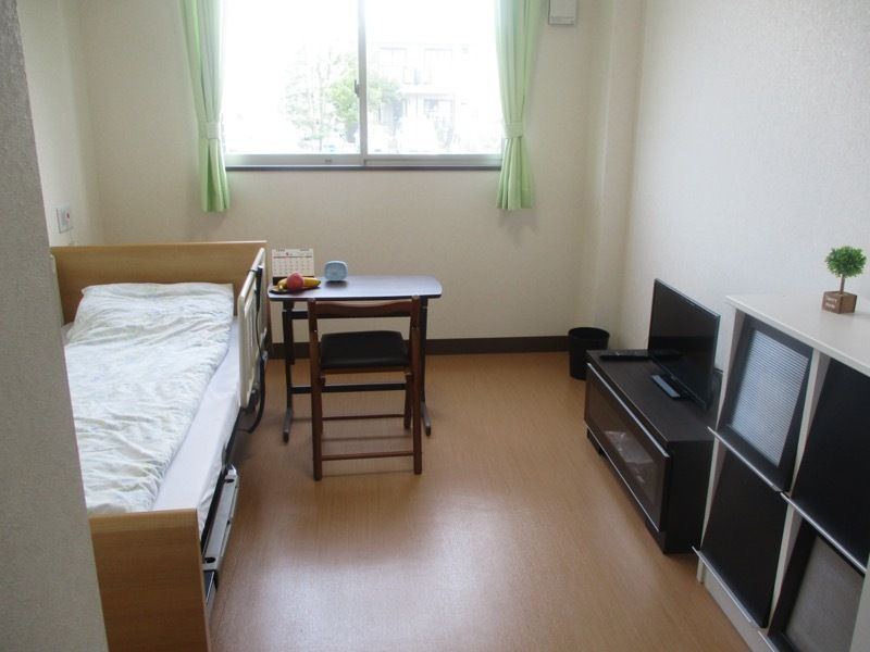カーテン付きの窓があり、テレビや寝具付きのベッドが置かれたフローリングの部屋。