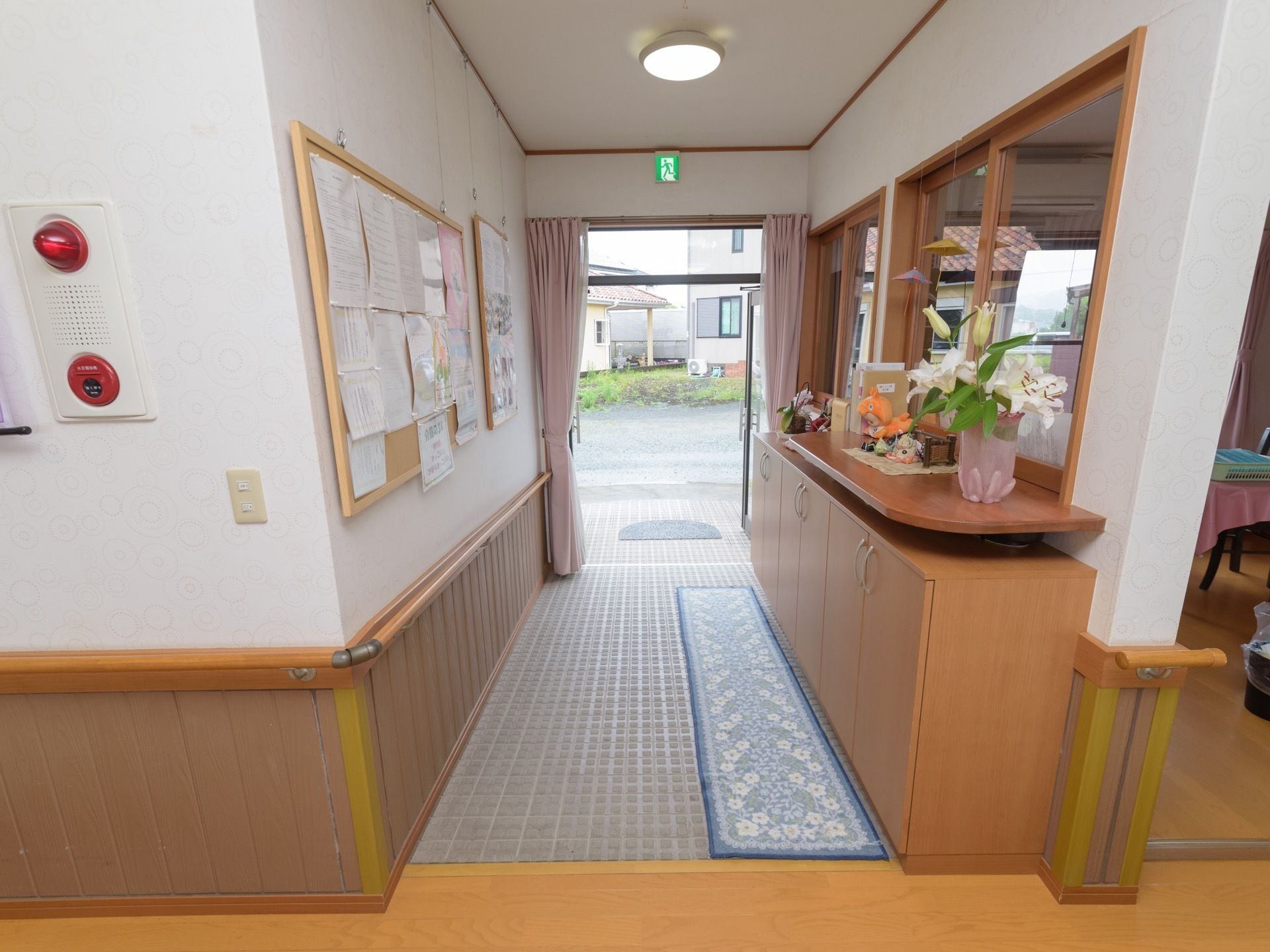 ピンク色のカーテンが掛けられた玄関があり、床はタイルになっている。壁にコルクボードが掛けられ、たくさんの紙が貼られている。