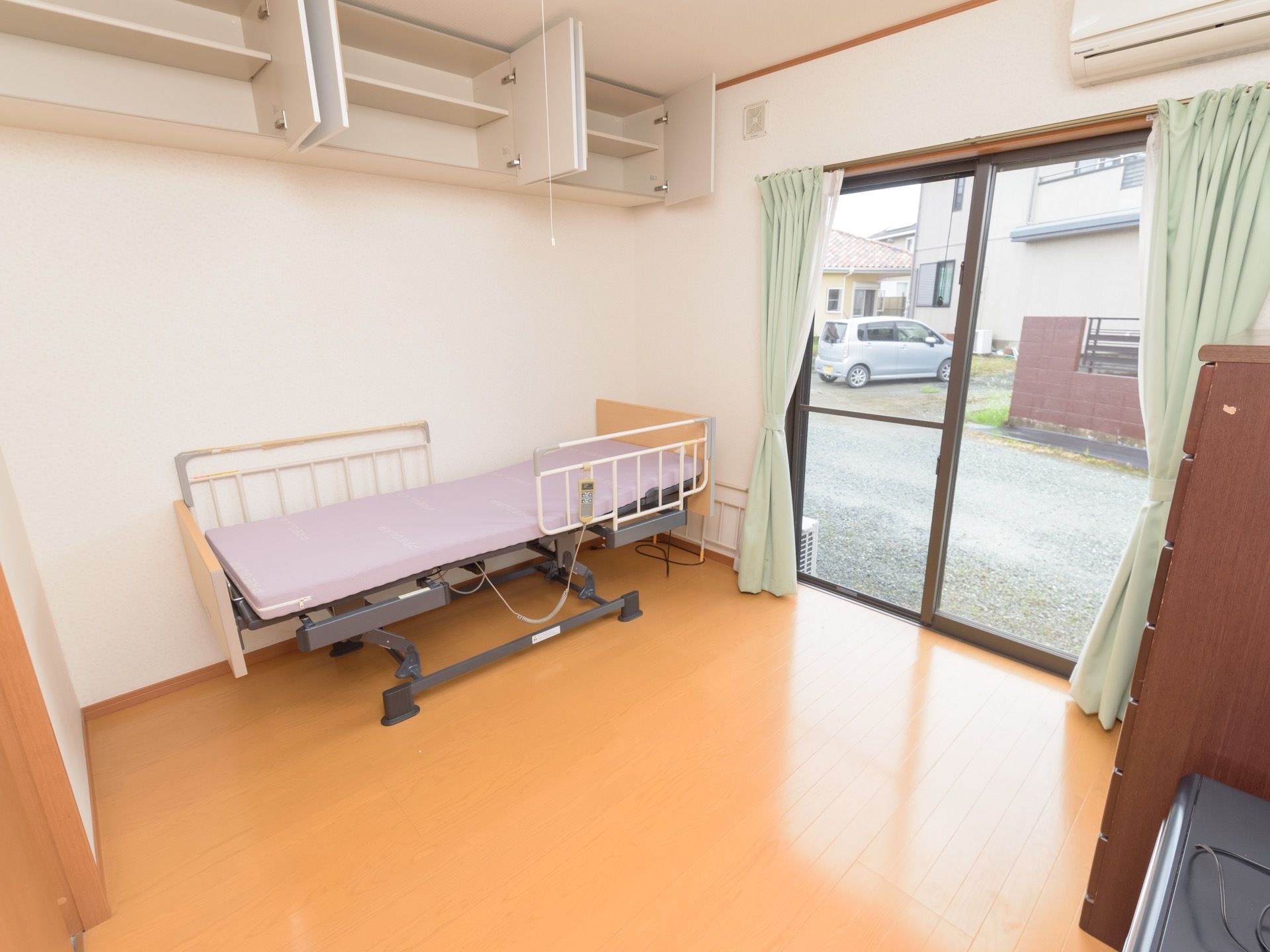 レースカーテンと薄いグリーンのカーテンが掛けられた掃き出し窓がある。部屋の角に介護ベッドが置かれ、その上に収納が設置されている。