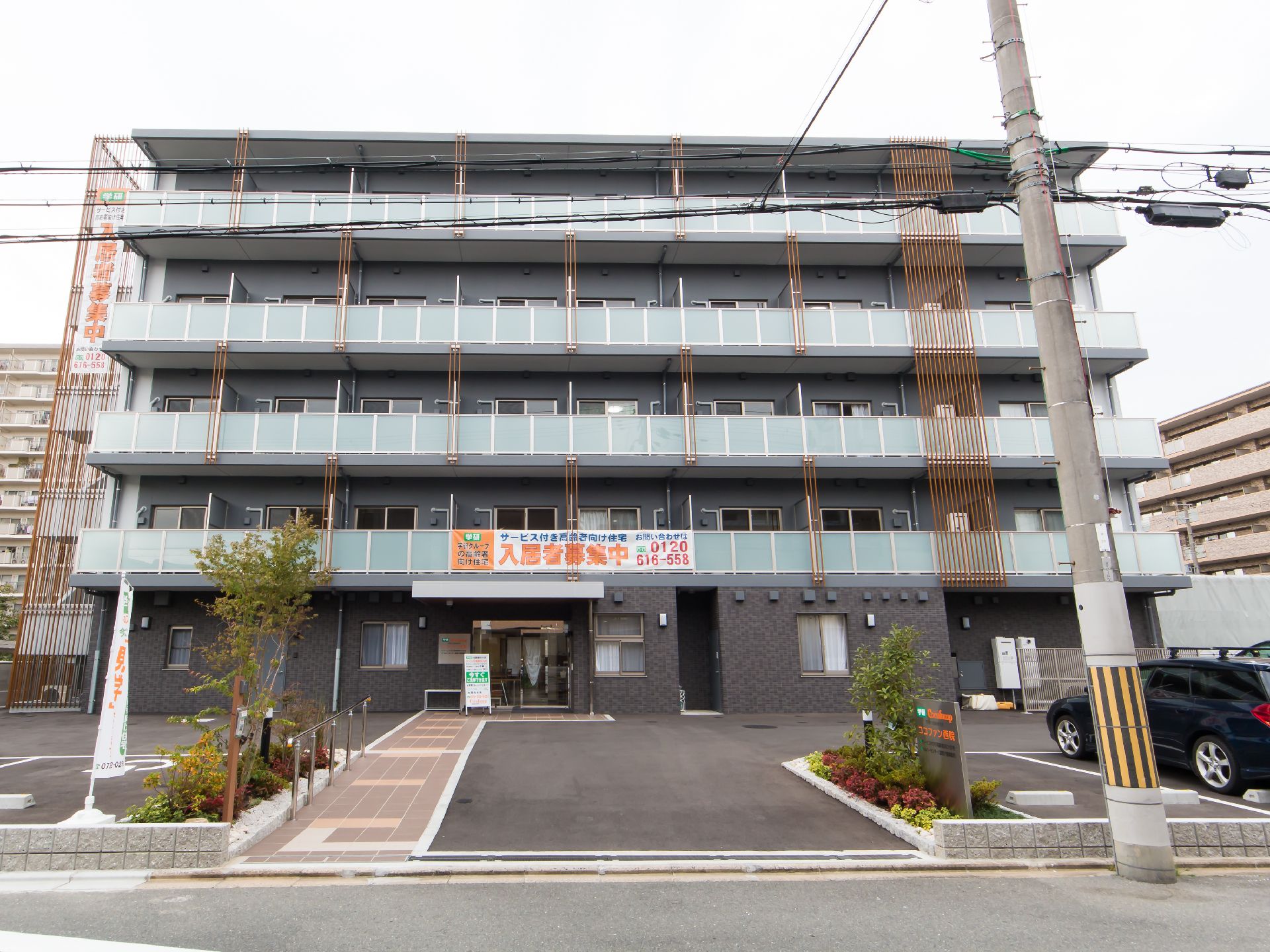 駐車場付きの５階建ての建物。各フロアにベランダがあり、玄関周辺には緑も植えられている。