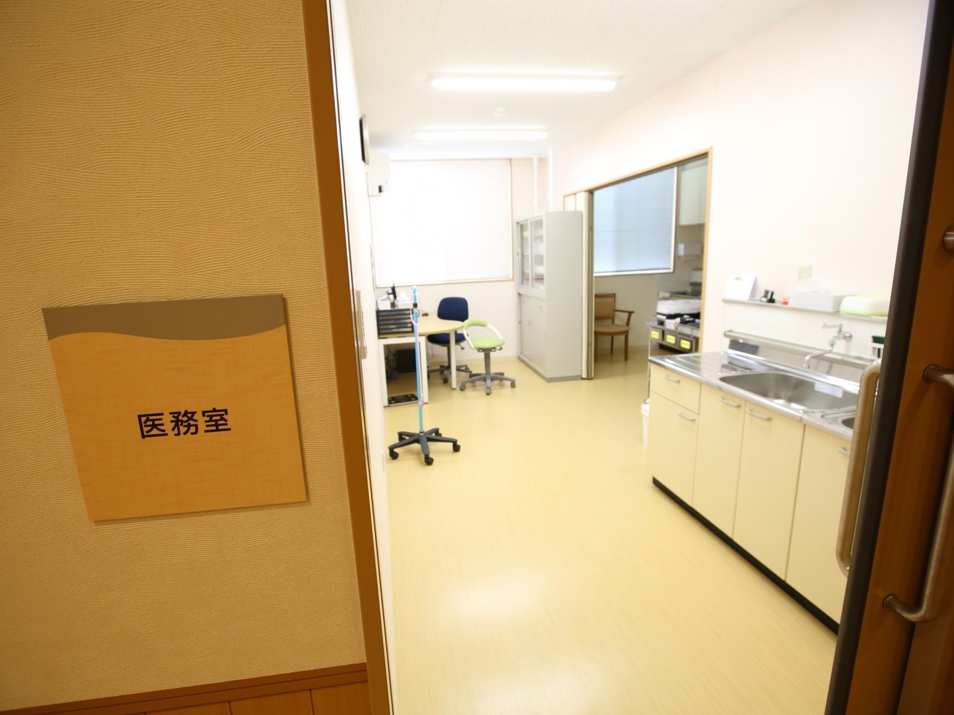 医務室と書かれた部屋には、テーブルや机、椅子、灰色の棚のほか、流し台も設置されている。また、奥にも部屋が続いている。