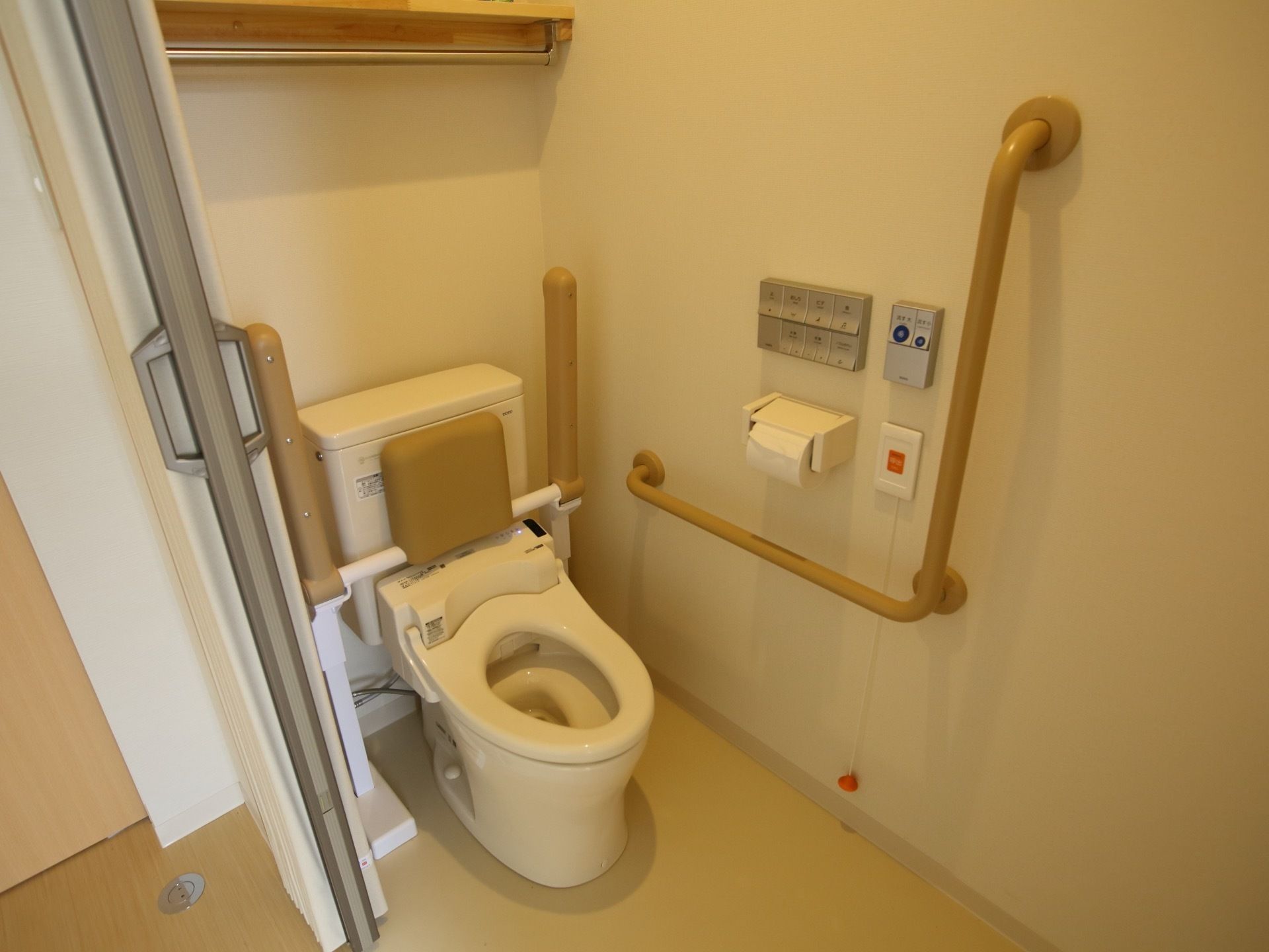トイレはアコーディオンカーテンで、床に段差がない。温水洗浄便器仕様で、背もたれや肘掛け、壁際に手すりがある。
