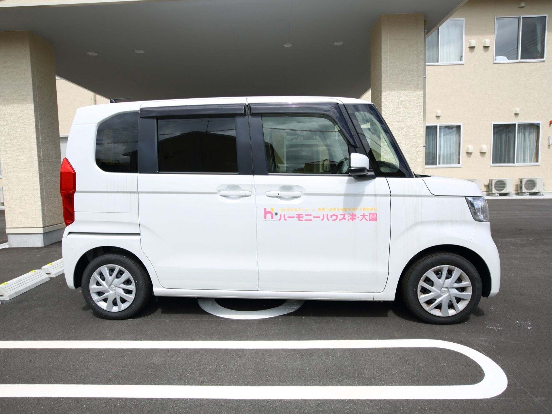 玄関前の駐車スペースに施設所有の自動車が停められている。白色のワゴンタイプで運転席のドアに施設名が書かれている。