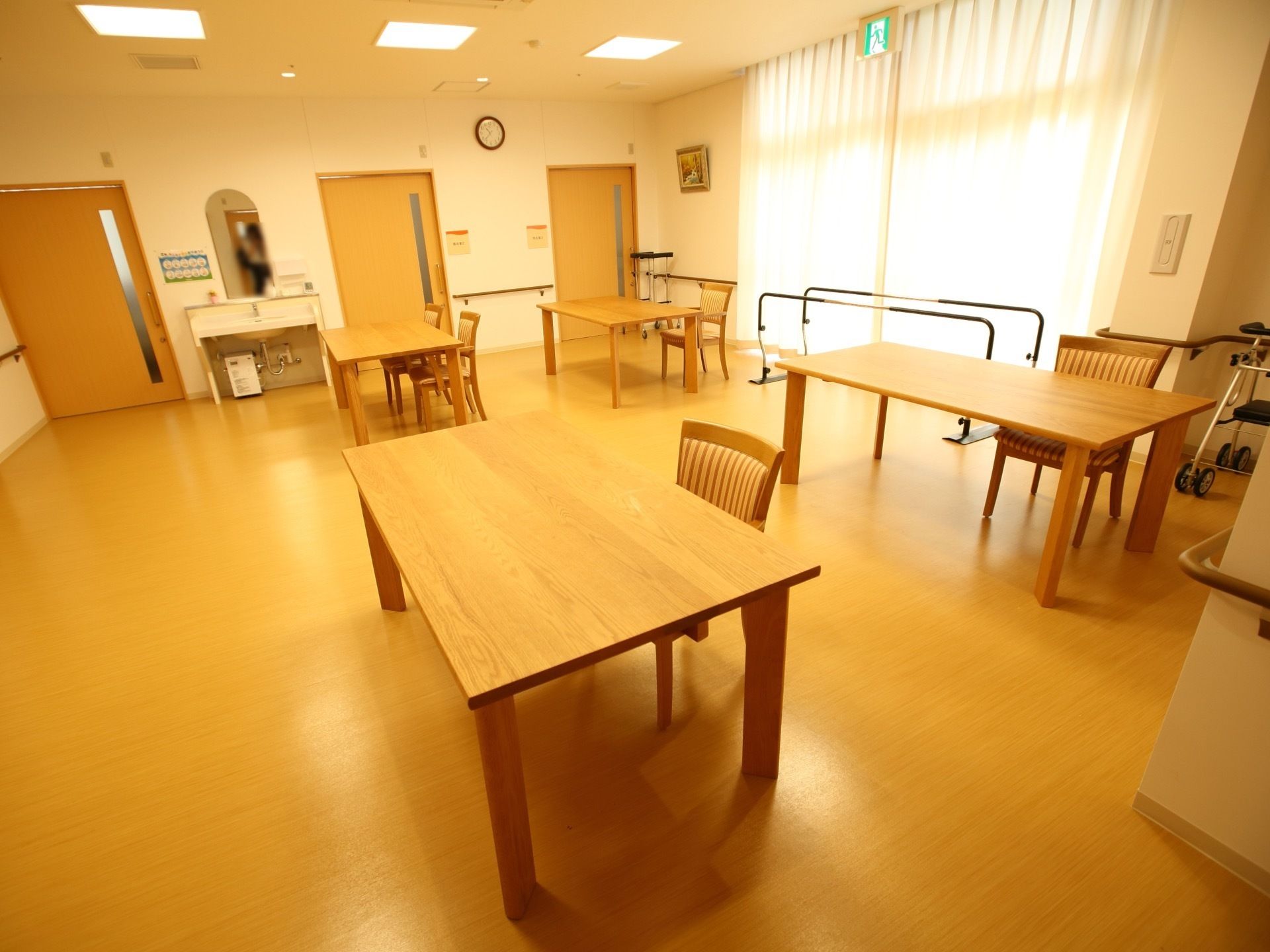 大きな窓が設置されている明るい共有スペース。複数の木製のテーブルや椅子が設置されているほか、洗面台や歩行器もある。