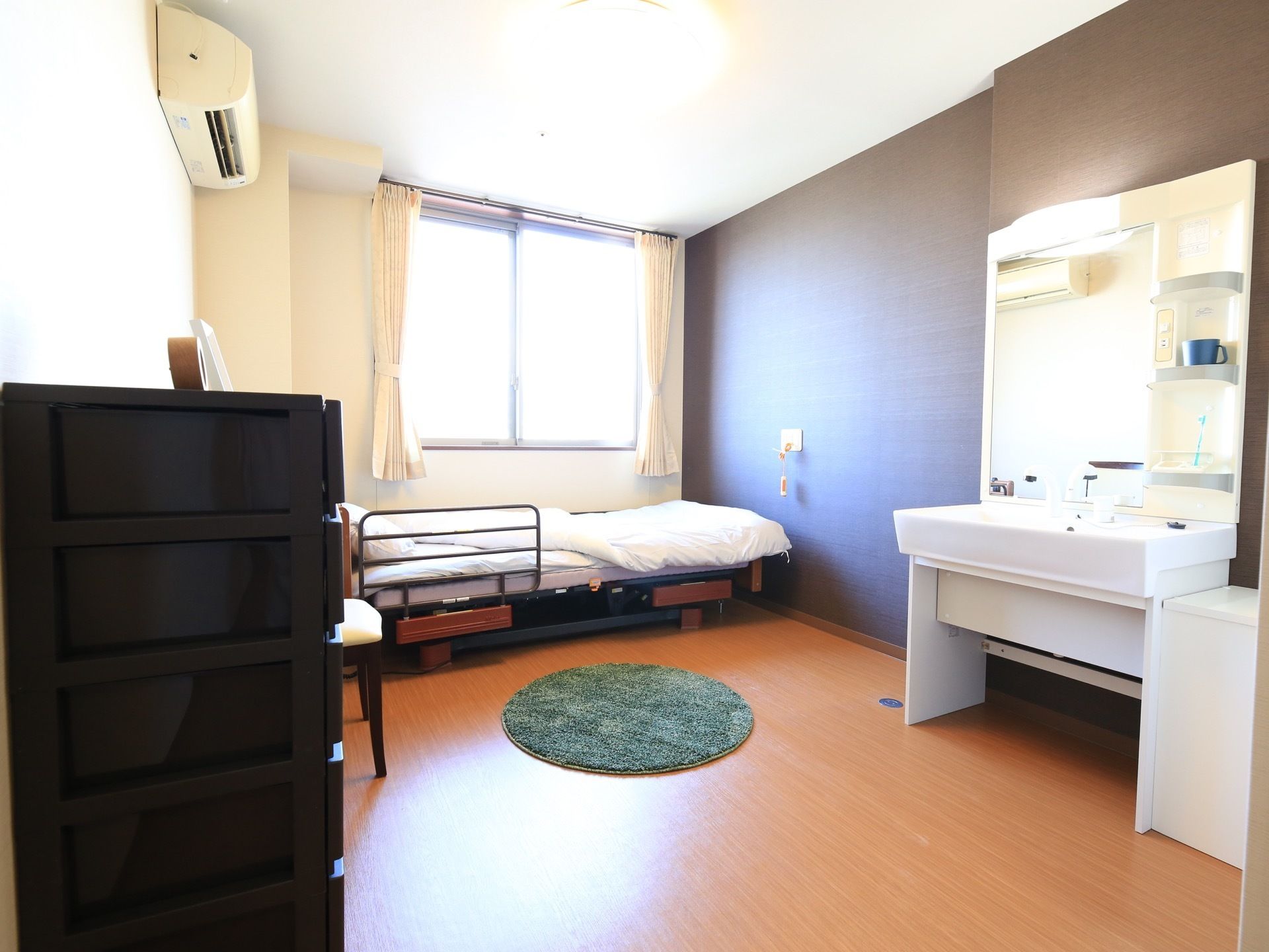 白い壁と一部が黒い壁になっている居室がある。カーテンの掛かった窓があり、側には介護用ベッドが置かれ、洗面台がある。