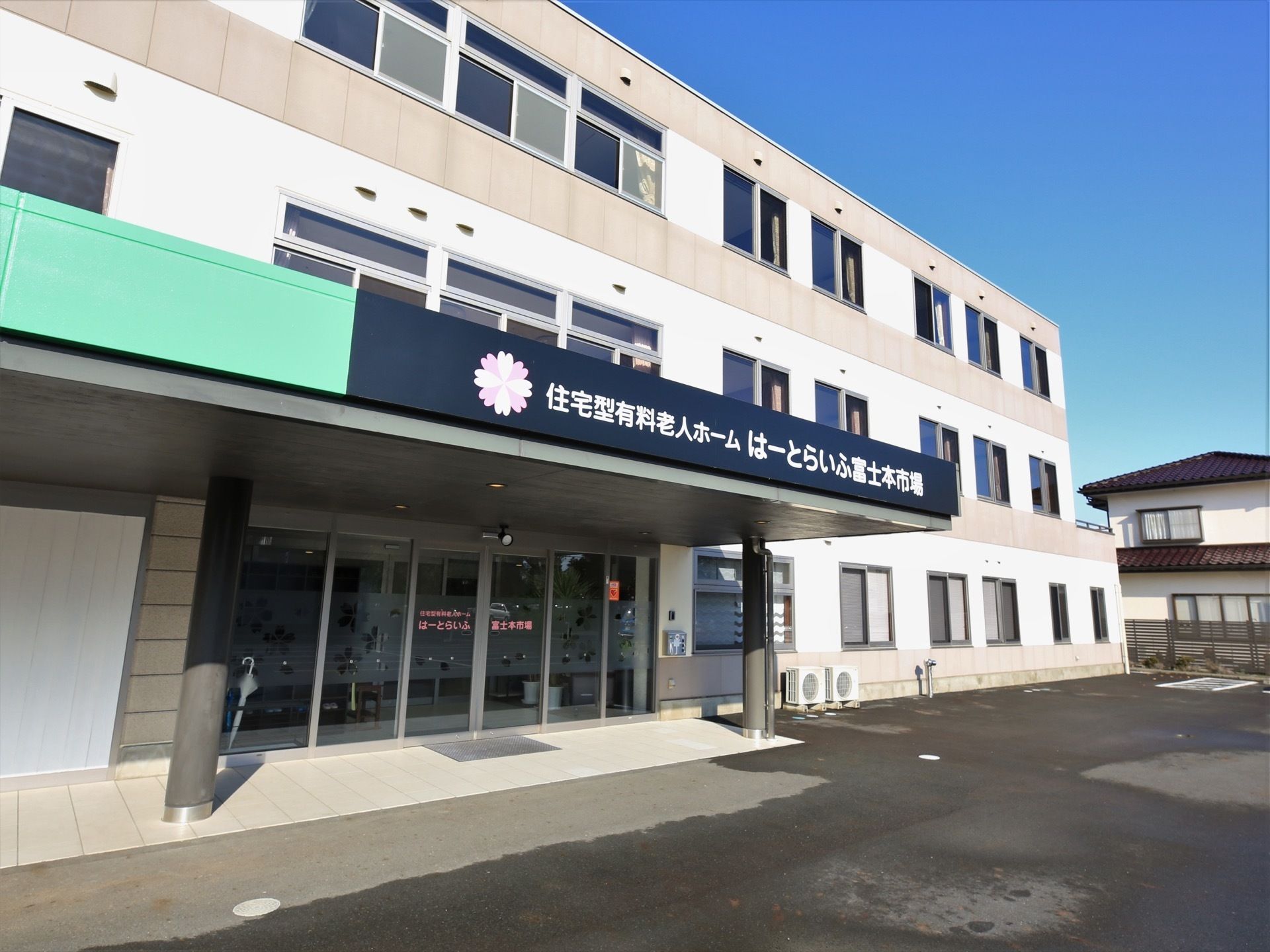 白い壁の３階建ての大きな建物がある。玄関の上には大きなルーフが付いており、側面に施設名が大きく書かれている。