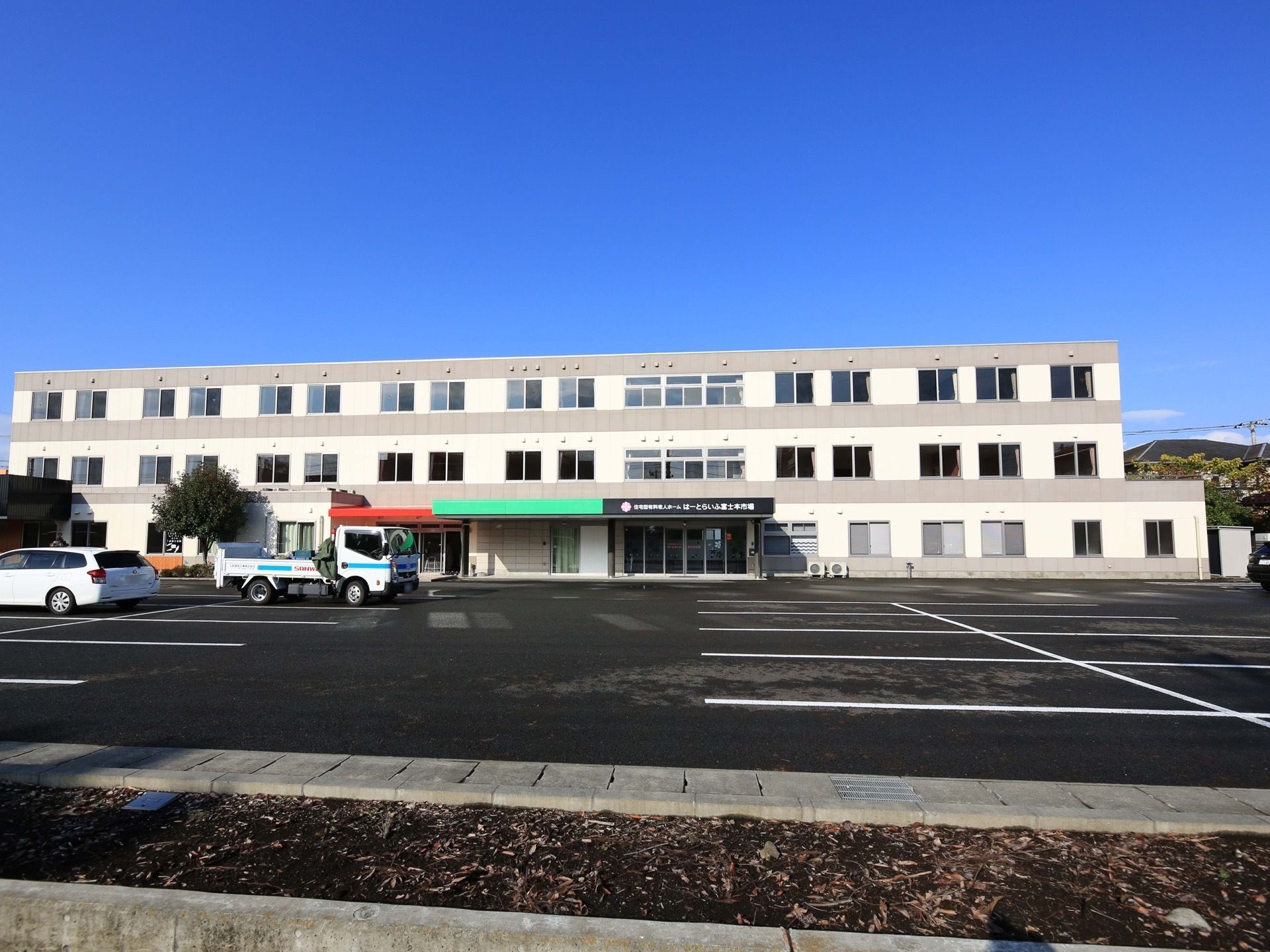 ３階建ての大きな建物があり、全面部分が広い駐車場になっている。地面は舗装され、白線で駐車スペースが描かれている。