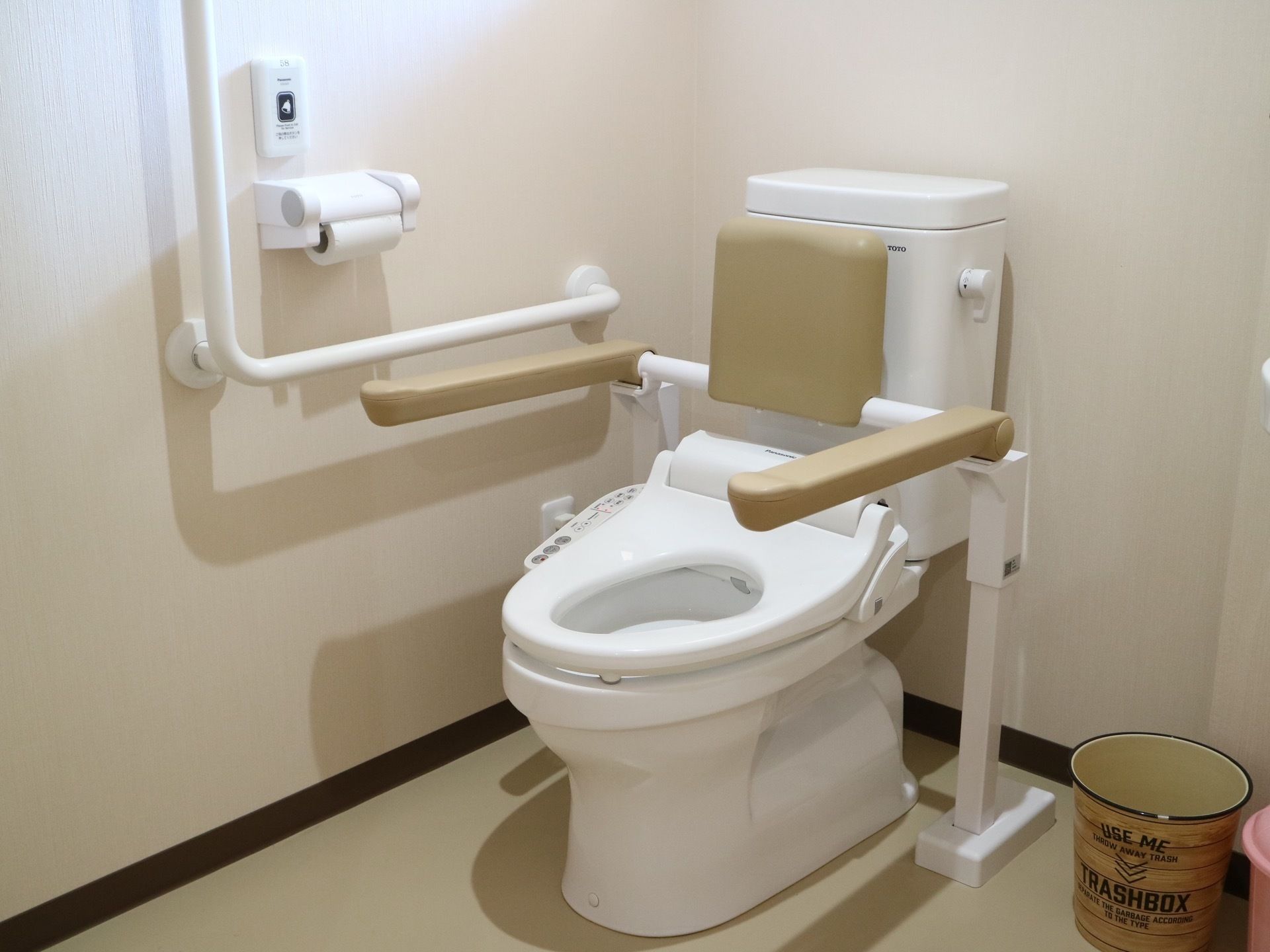 トイレの便座の周囲には手すりと背もたれがある。また壁にはＬ字型の手すりとトイレットペーパーホルダー、ナースコールが設置されている。