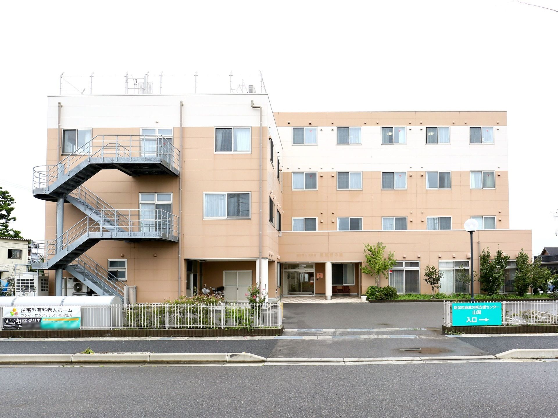 ベージュとホワイトの落ち着いたカラーの施設外観である。4階建ての建物である。敷地内には植栽が施されている。