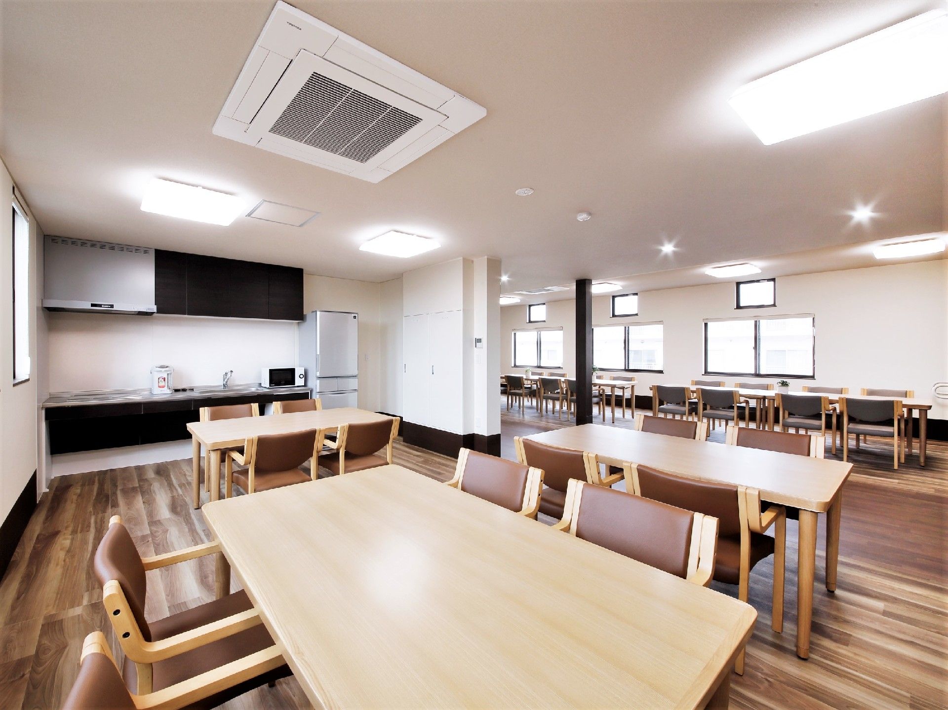 アイボリーのテーブルと茶色い座面の椅子が置かれた部屋。広いフロアの中にいくつも置かれている
