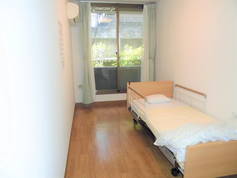 寝具付きのベッドが置かれたフローリングの部屋にはカーテン付きの窓があり、エアコンもある。
