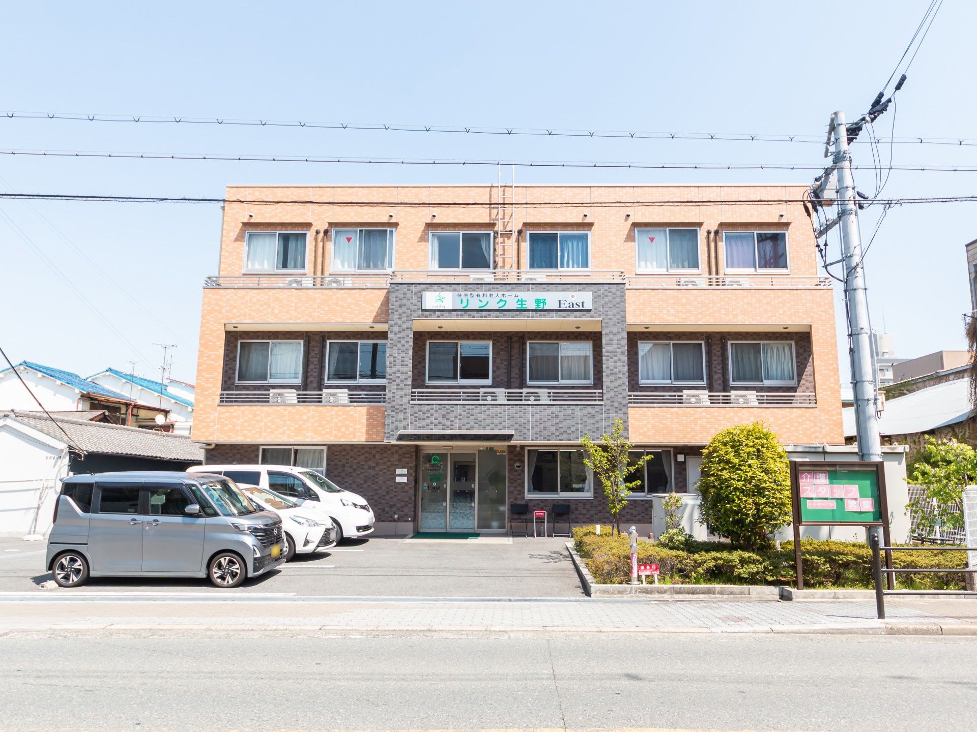 ブラウン系の外壁の3階建ての建物の前には駐車場があり、日当たりのいい場所に建っている。