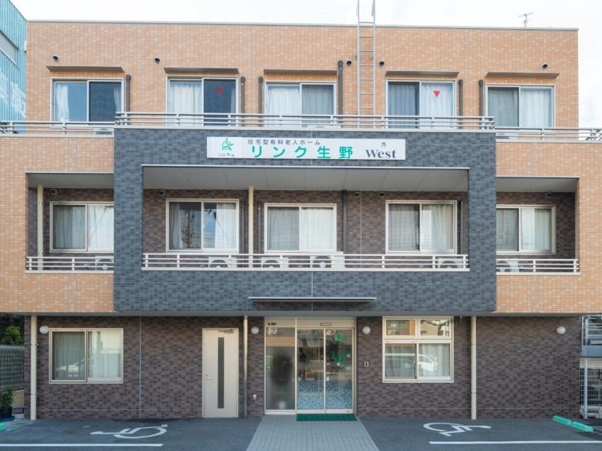 ブラウン系の外壁の3階建ての建物の前には駐車場があり、各フロアにベランダもある。