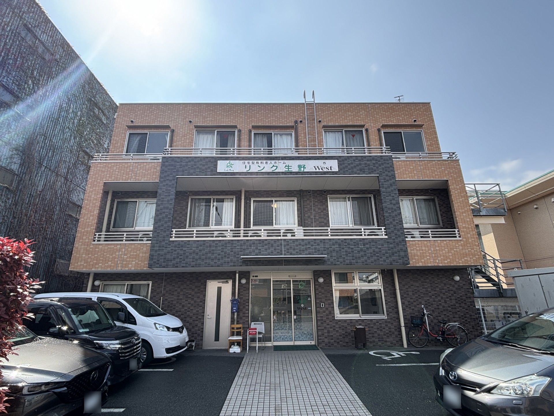 ブラウン系の外壁の各フロアにベランダもある3階建ての建物の前には駐車場がある。