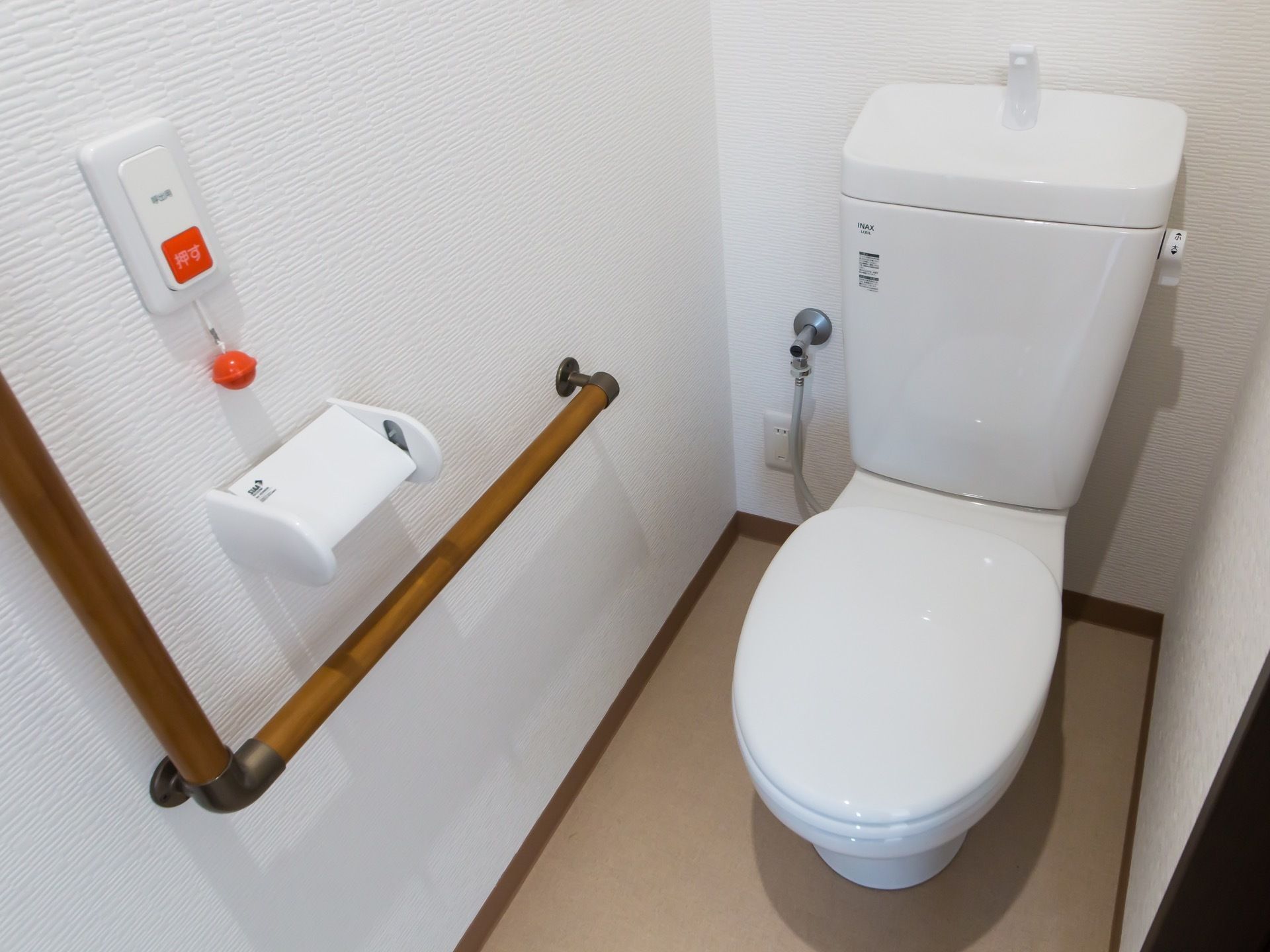真っ白い壁と便器が清潔さを感じるトイレである。壁に木製のL字型手すりとペーパーホルダー、ナースコールが取り付けてある。