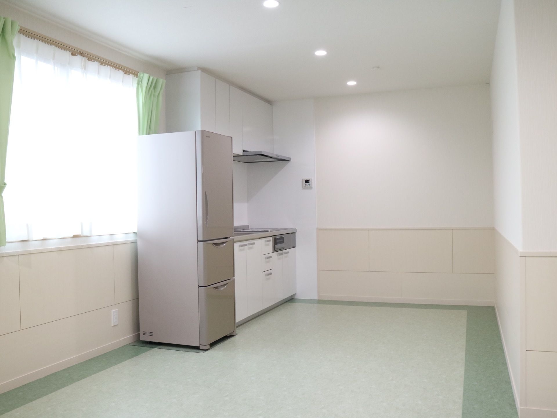 キッチンが設置されており、その左側に冷蔵庫が置かれている。腰掛け窓があり、レースと緑のカーテンが掛けられている。