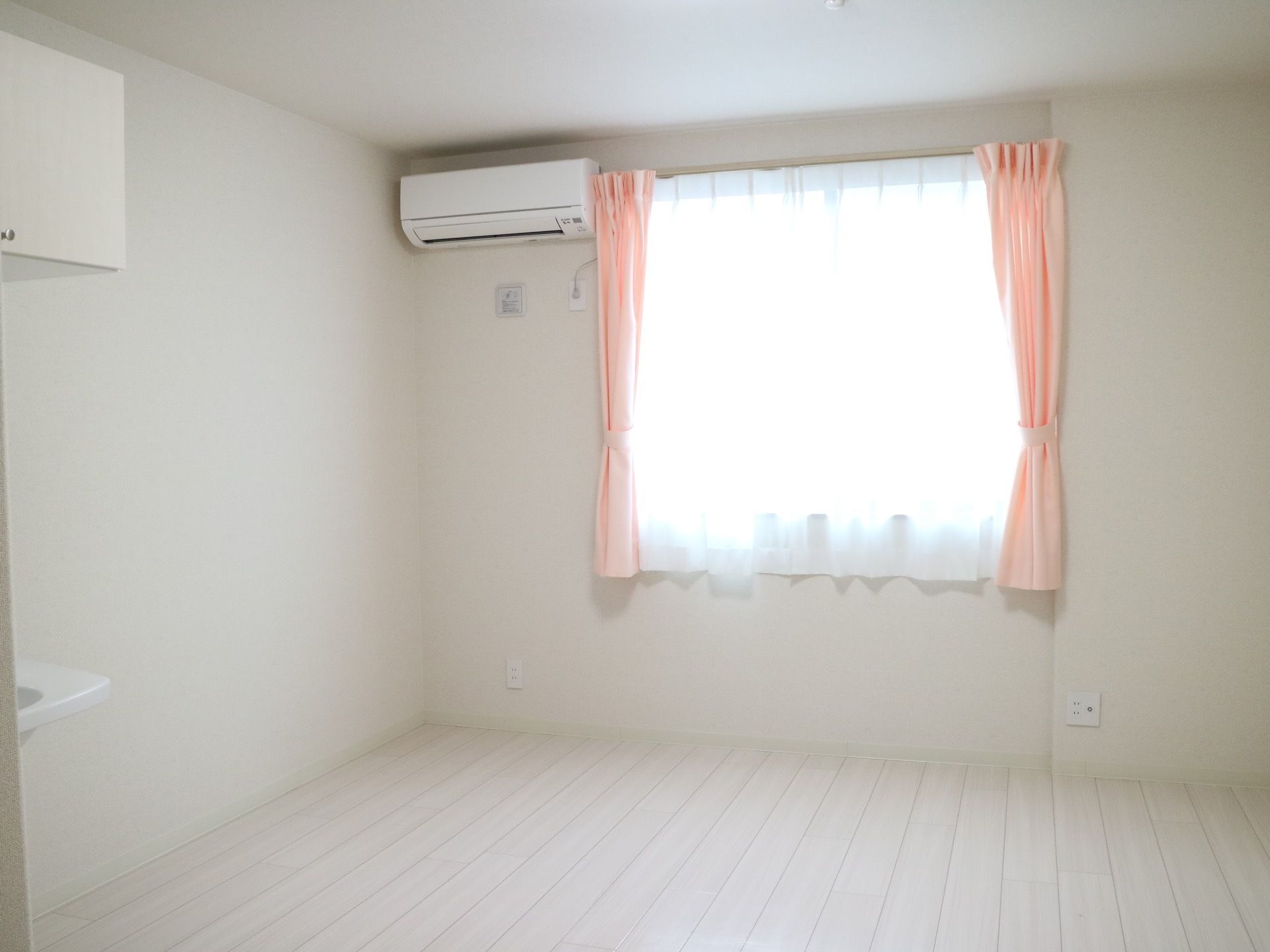 全体が白に統一された清潔感のある部屋である。腰高窓があり、ピンクのカーテンが付けられている床はフローリングになっている。