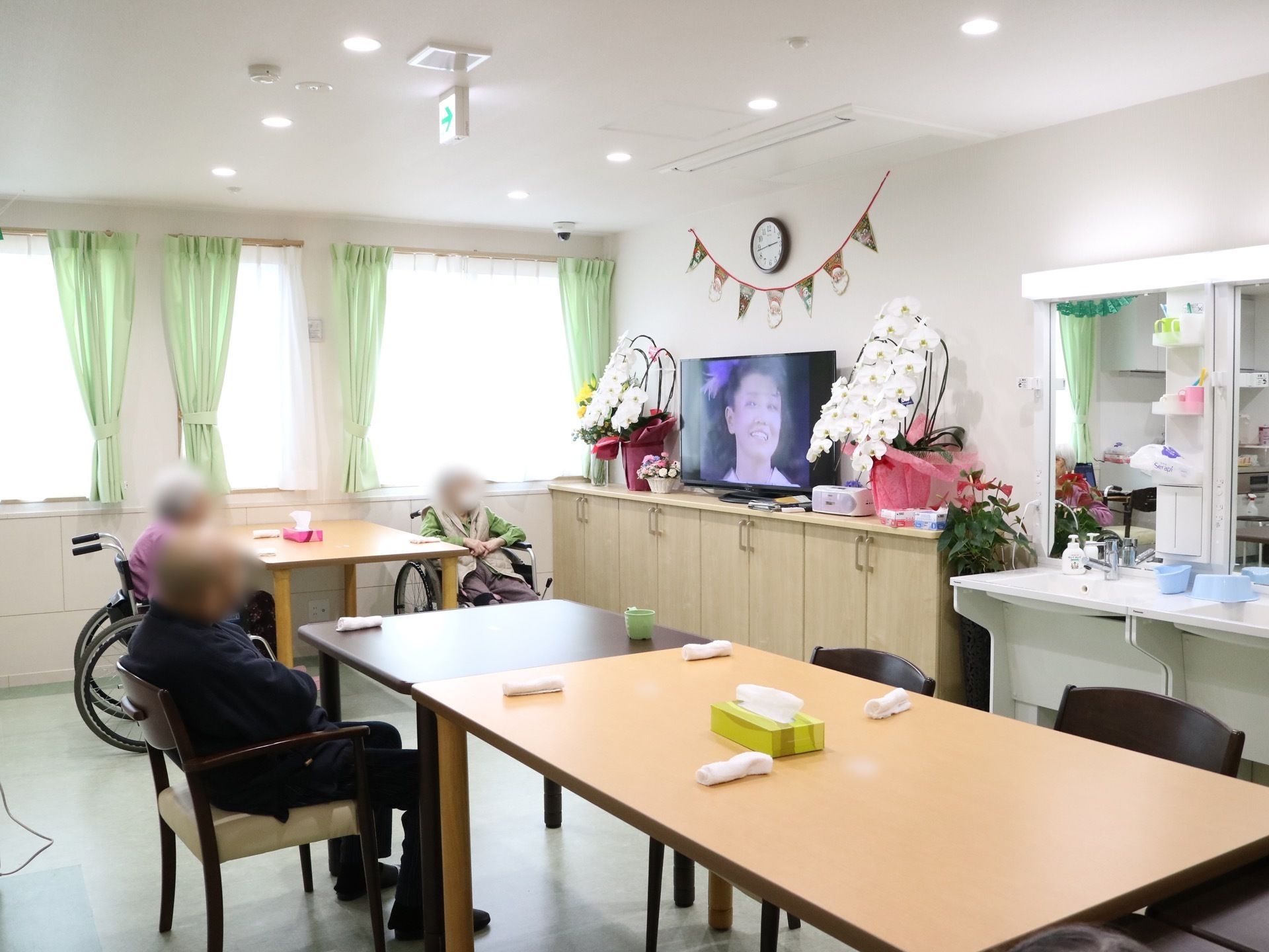 共有スペースに入居者様が３名集まって、演歌歌手が映っているテレビを見ている。テレビの両脇にきれいな花が飾られている。