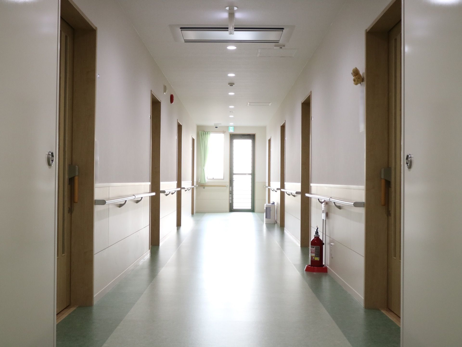 廊下のつきあたりに、扉と腰高の窓がある。廊下の壁際に消火栓が置かれている。非常ベルが壁の上の方に設置されている。