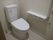 トイレにきれいな白色の便座が設置されている。壁に取り付けられた棚板の隣に赤色の呼び出しボタンがある。