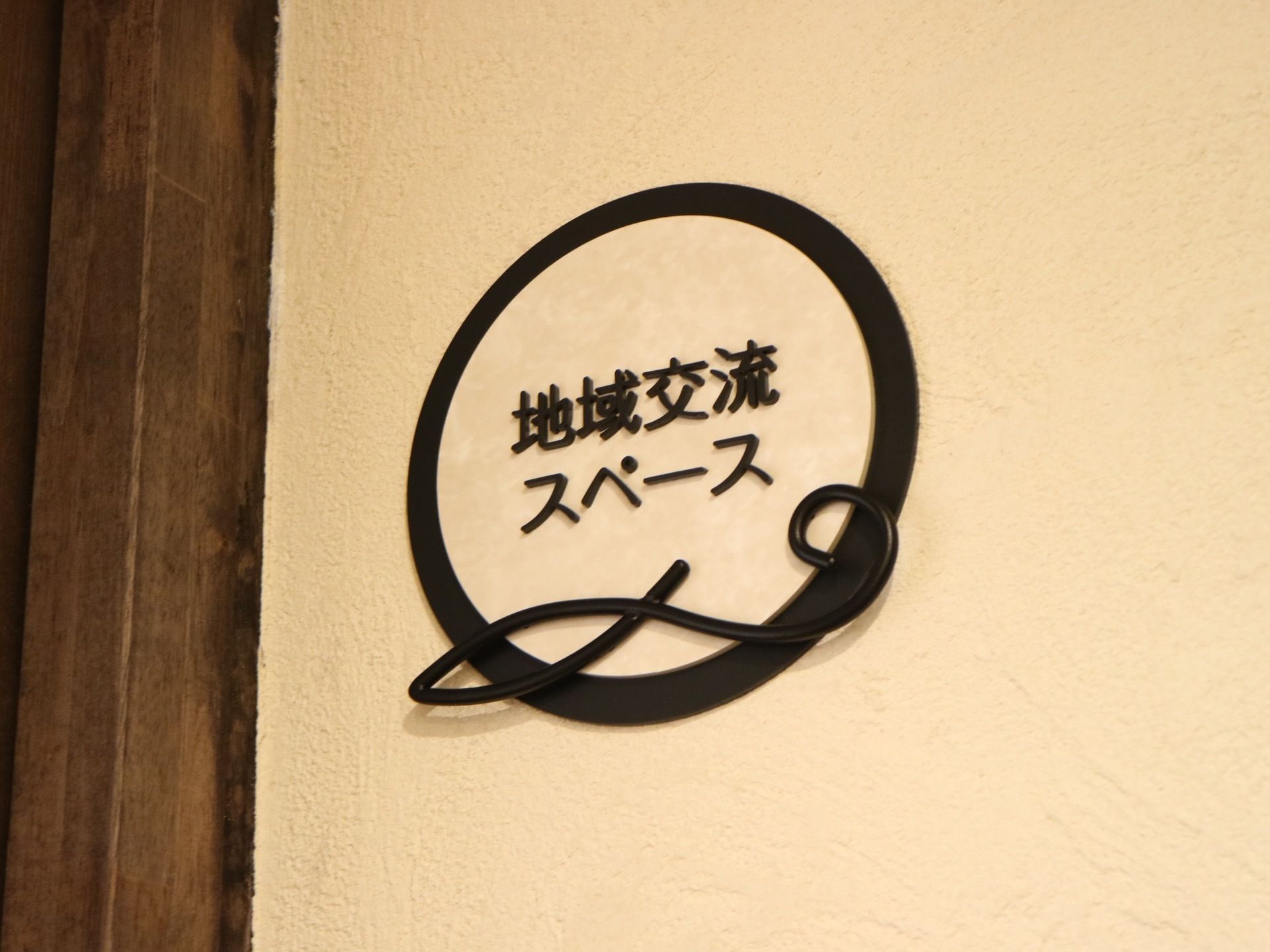 館内にある地域交流スペースの看板である。白い壁の中央に黒い素材を使用して作られたものが取り付けられている。