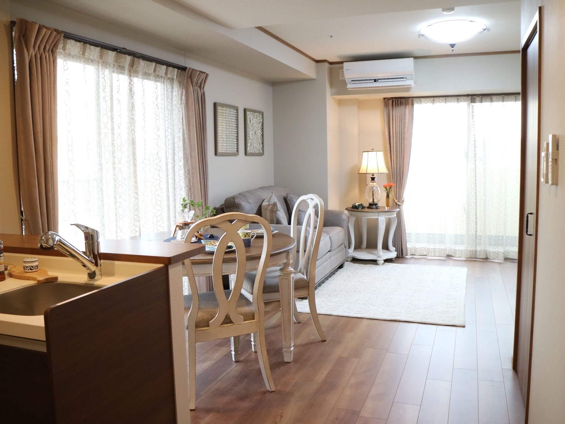 左手前にキッチンの流しが見えている。奥に広間が見えており、２ヵ所に窓がある。円卓とソファが用意されている。