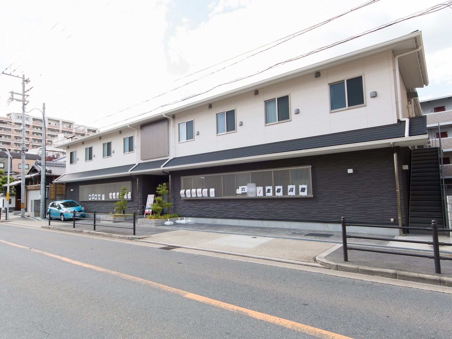 道路から見た施設建物。車通りの多い大きな通りに接しており、前面の敷地には縦列専用の駐車スペースが設けられている。