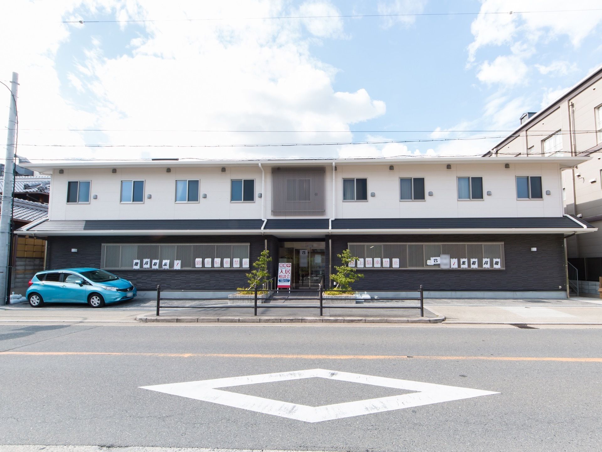 正面から見た施設建物の様子。１階と２階で外壁の色とデザインが違っている。敷地には縦列専用の駐車スペースがある。