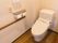 濃い色のフローリングの床の個室に洋式トイレがあり、壁には手すりとナースコール、自動洗浄器の操作パネルが設置されている。