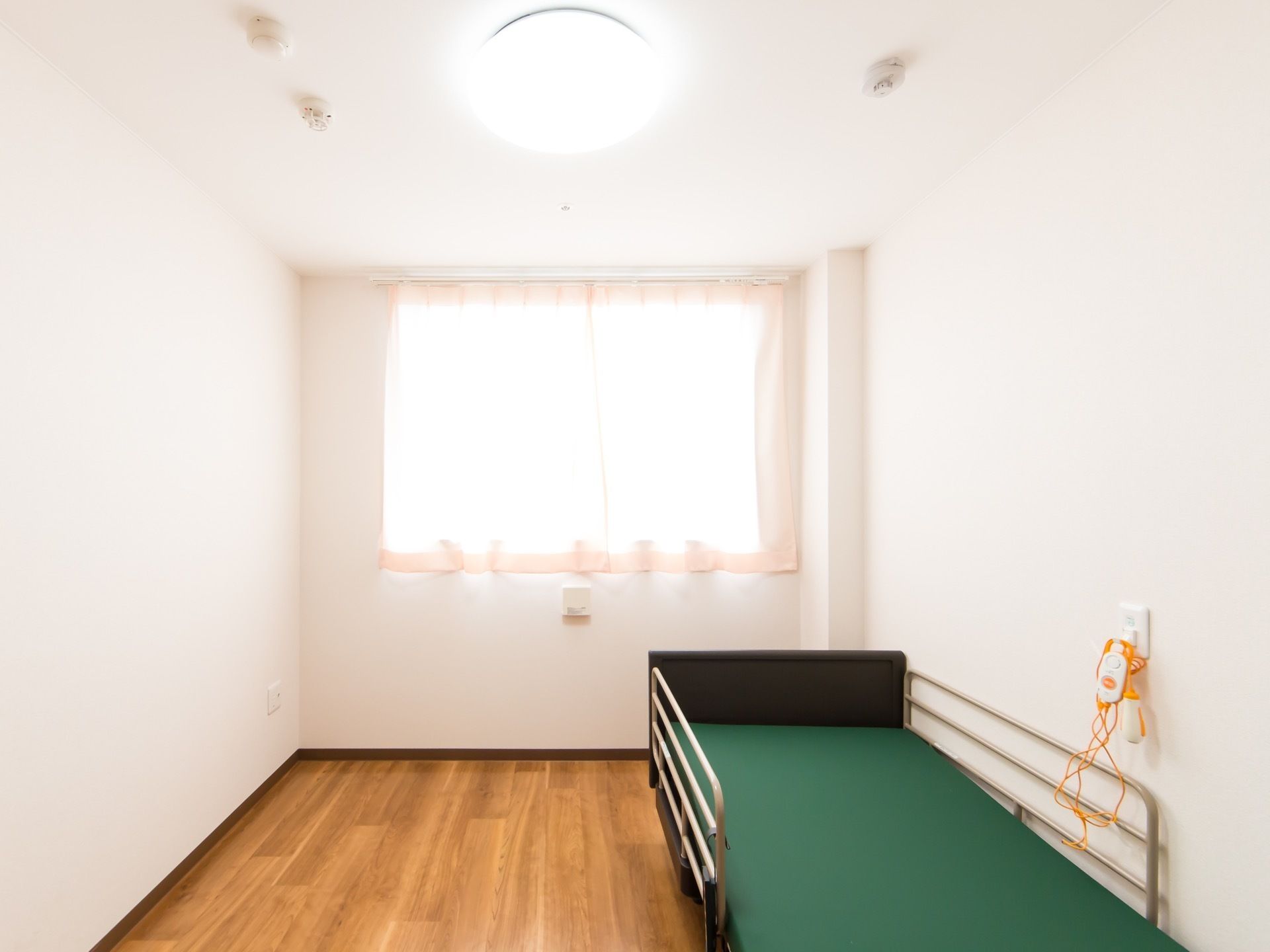 フローリングの床に白い壁の部屋で、右側にベッドが一台置かれている。ベッド脇の壁にナースコールが設置されている。