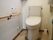 白い温水洗浄便座器が完備されたトイレである。壁には木製の手すりが設置され、コーナーには清掃用具が置かれている。