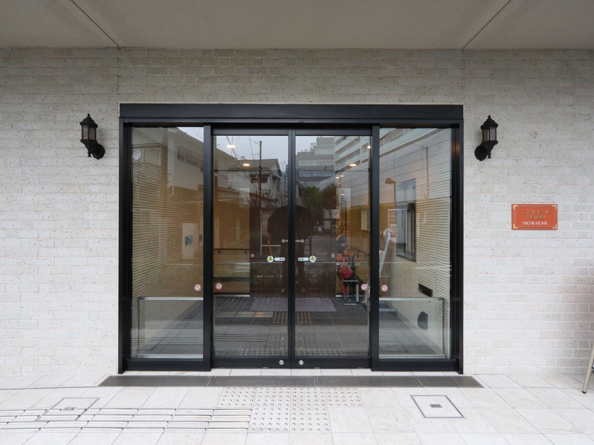 黒い縁取りのガラスの自動扉がある。扉の両側の壁に門灯が設置され、施設名が表示されたオレンジ色のパネルが取り付けられている。