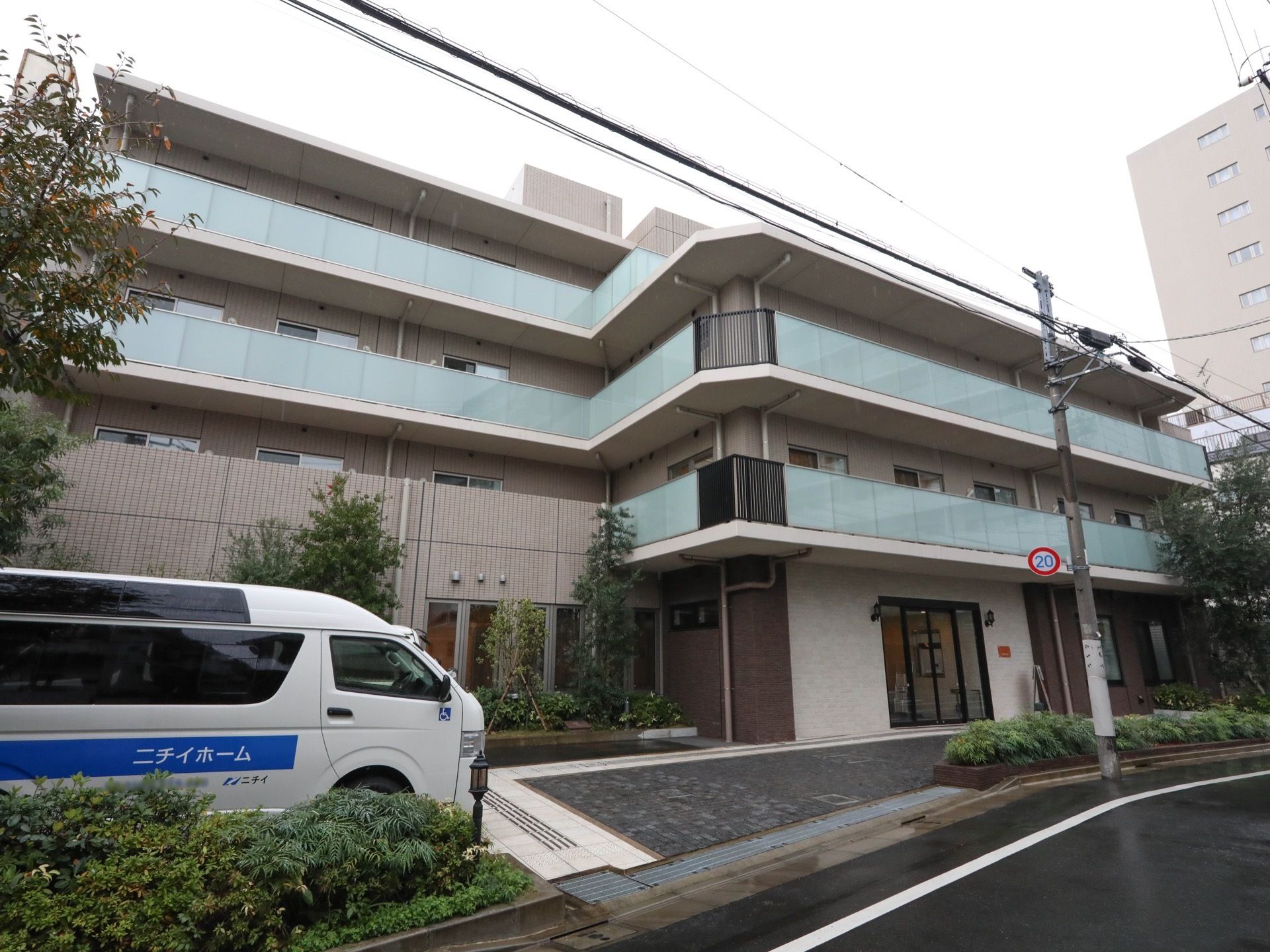 ４階建ての建物のベランダにはパネルタイプの柵が設置されている。植栽スペースの奥に施設名が表示された車が停められている。
