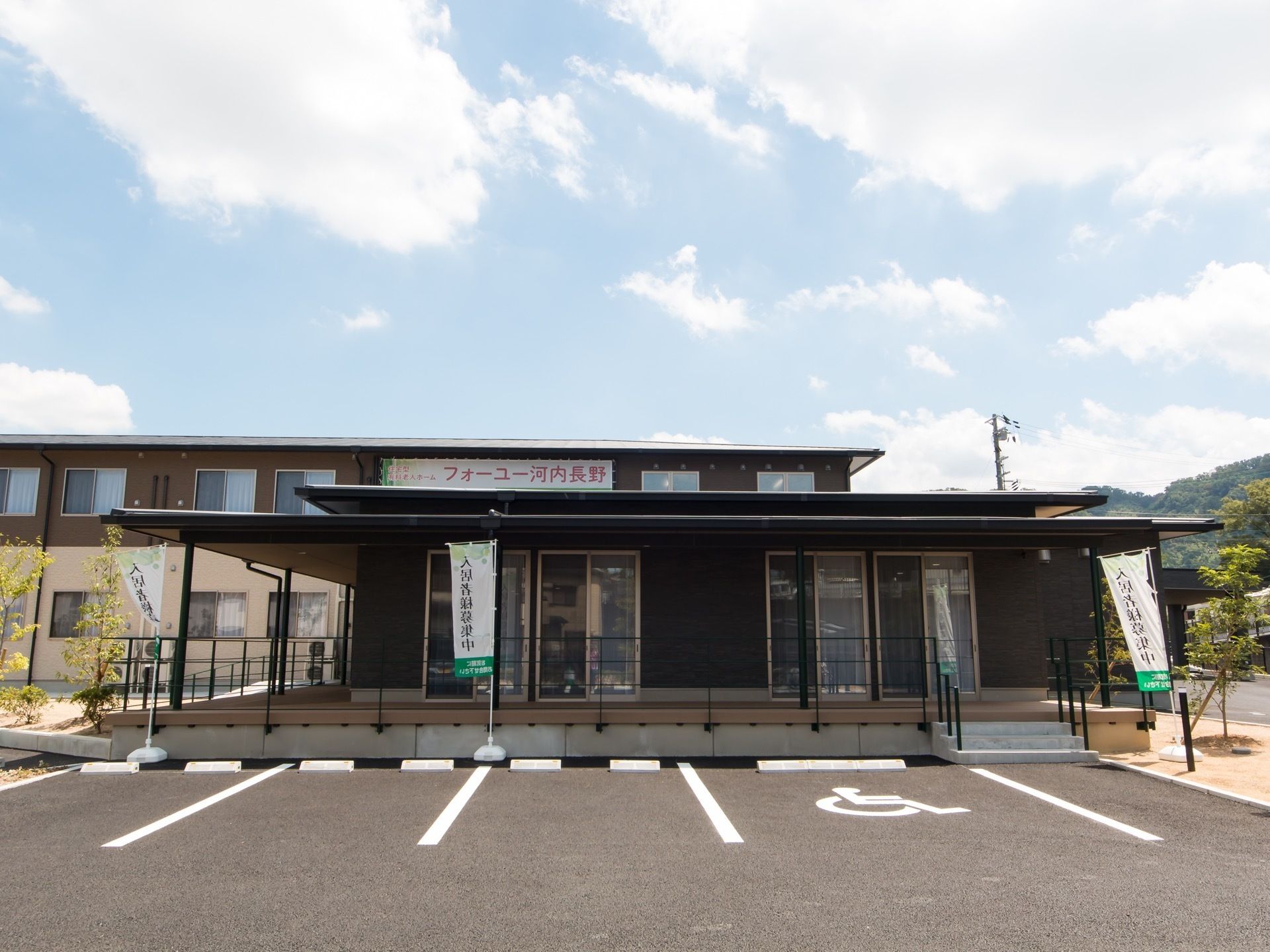 2階建の建物の前にウッドデッキを備えた平屋の建物が建っている。車椅子を使う方優先の駐車場が備わっている。
