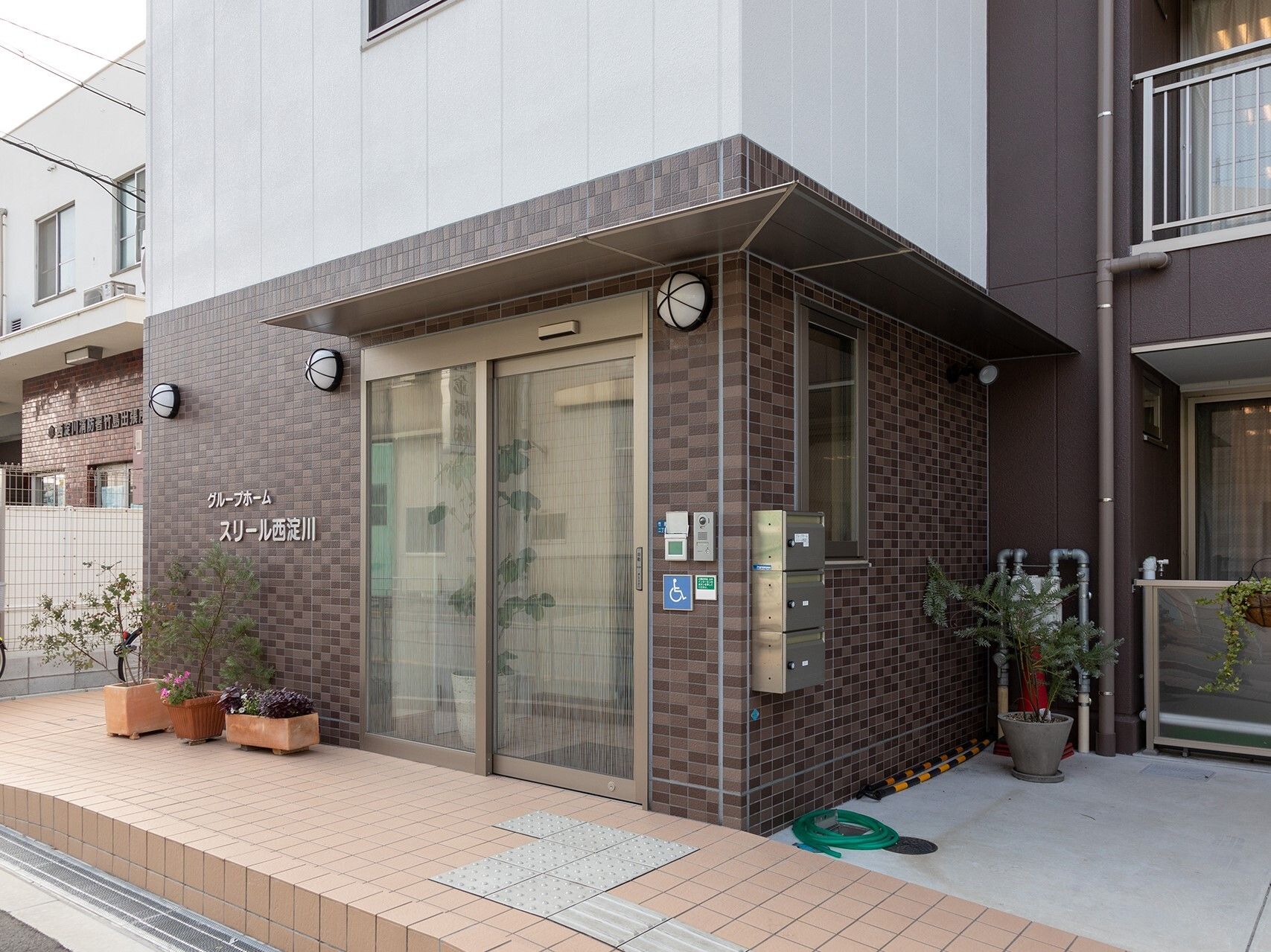 薬を飲もうとしている高齢女性のそばにはエプロンを付けた若い女性スタッフがいて、水の入ったコップを持っている。