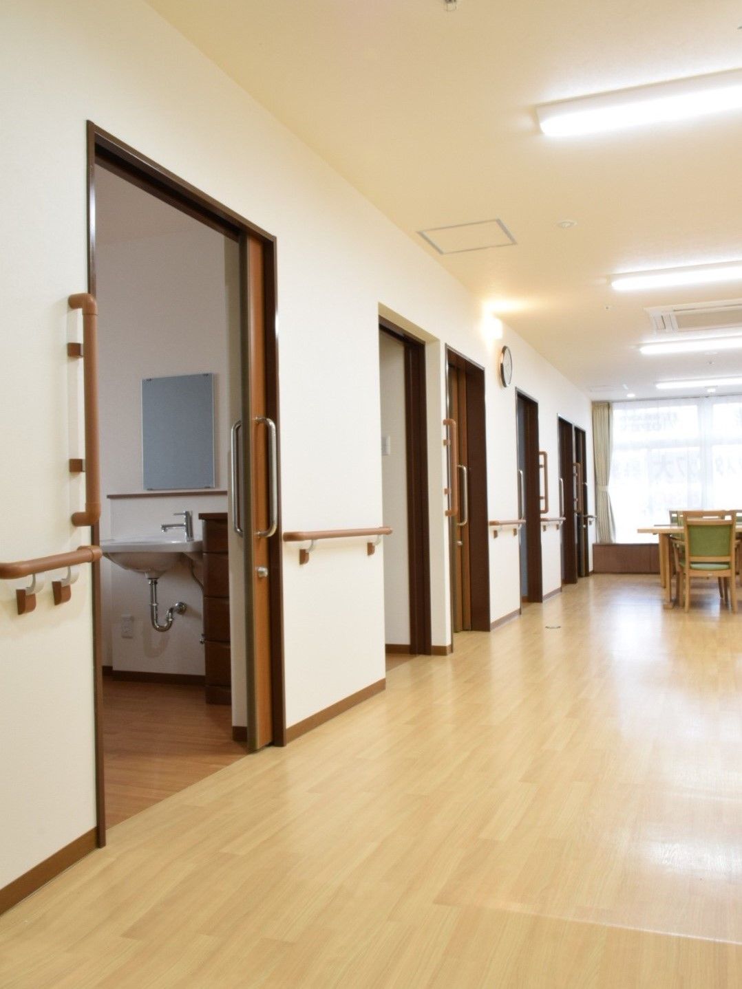 手すりが付いたまっすぐな廊下。奥にはカーテン付きの窓があり、居室には木製の引き戸が付いている。