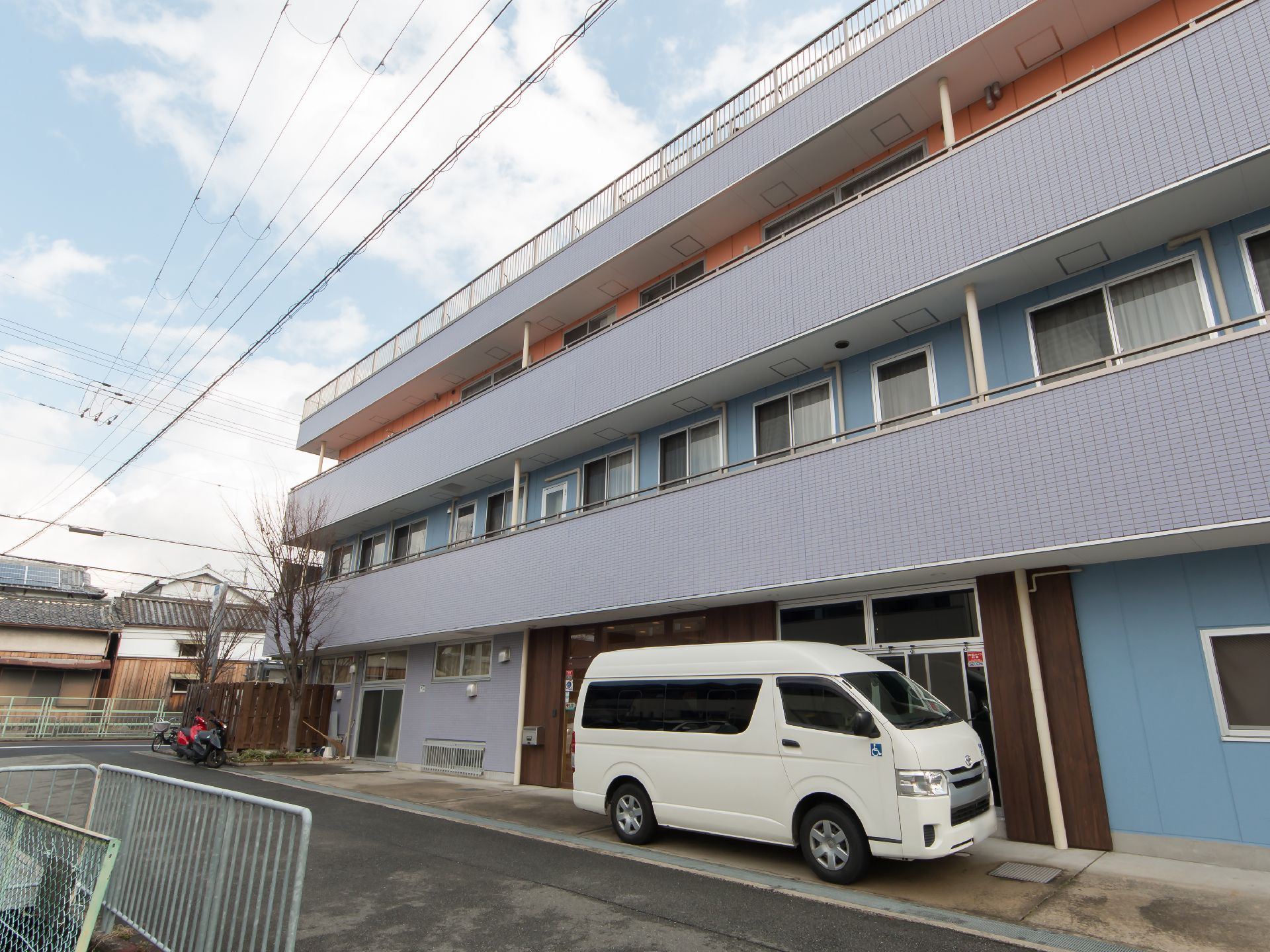 平屋根の３階建ての建物で、外壁の色が各階によって違うカラフルなデザインである。玄関前のスペースには施設所有の大型車が停められている。