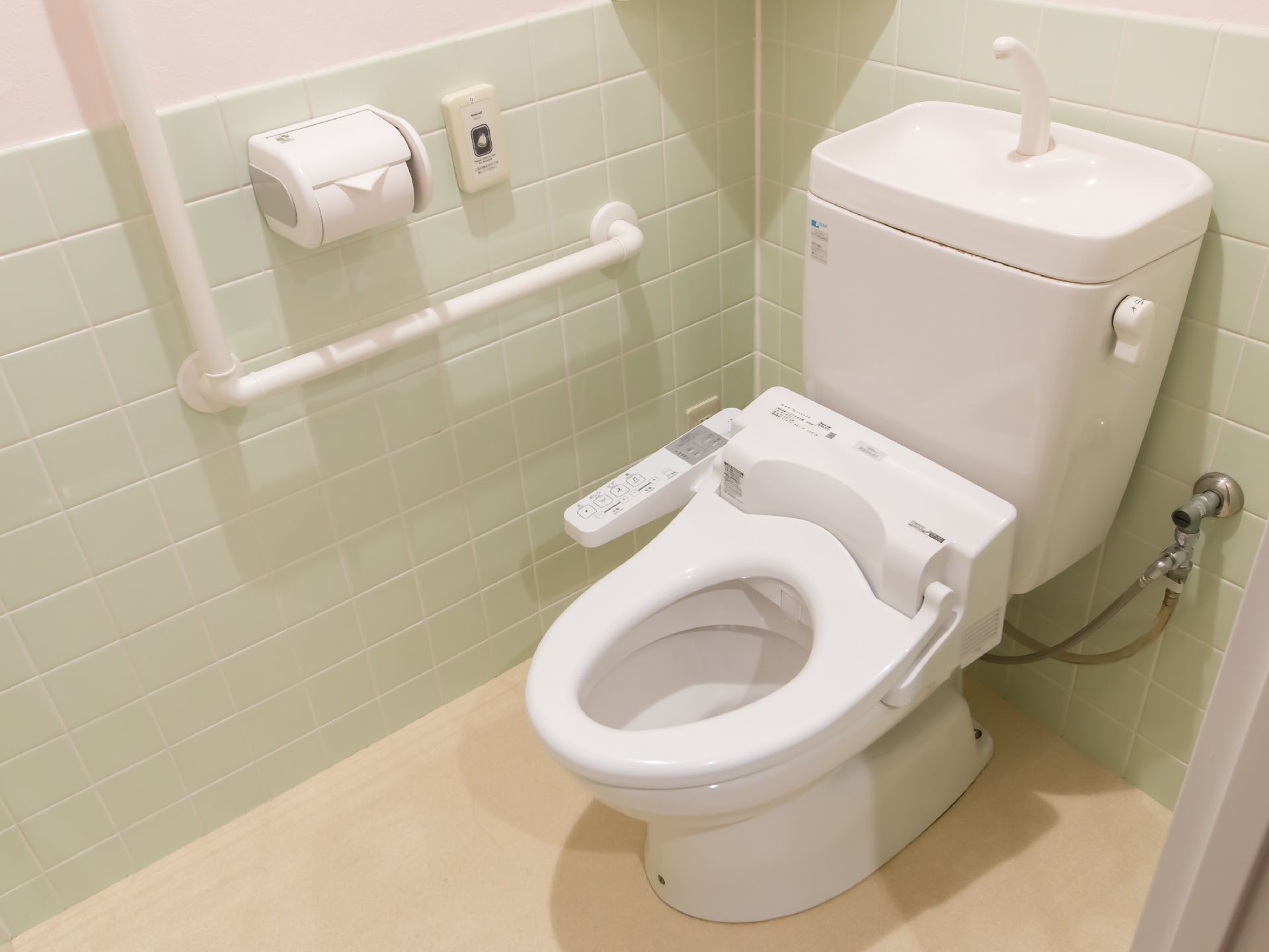 トイレは全体的に淡い色合いの個室である。蓋がない温水洗浄便座で、壁には手すりやナースコールが設置されている。