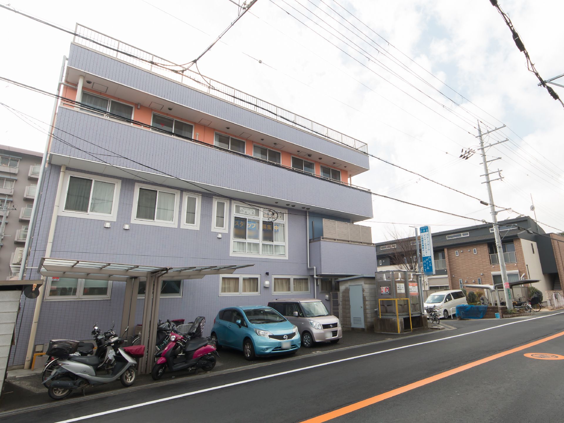 片側一車線の道路に面した３階建ての建物で、外壁はグレーとオレンジ色である。道路沿いに駐車スペースがあり、車が停められている。
