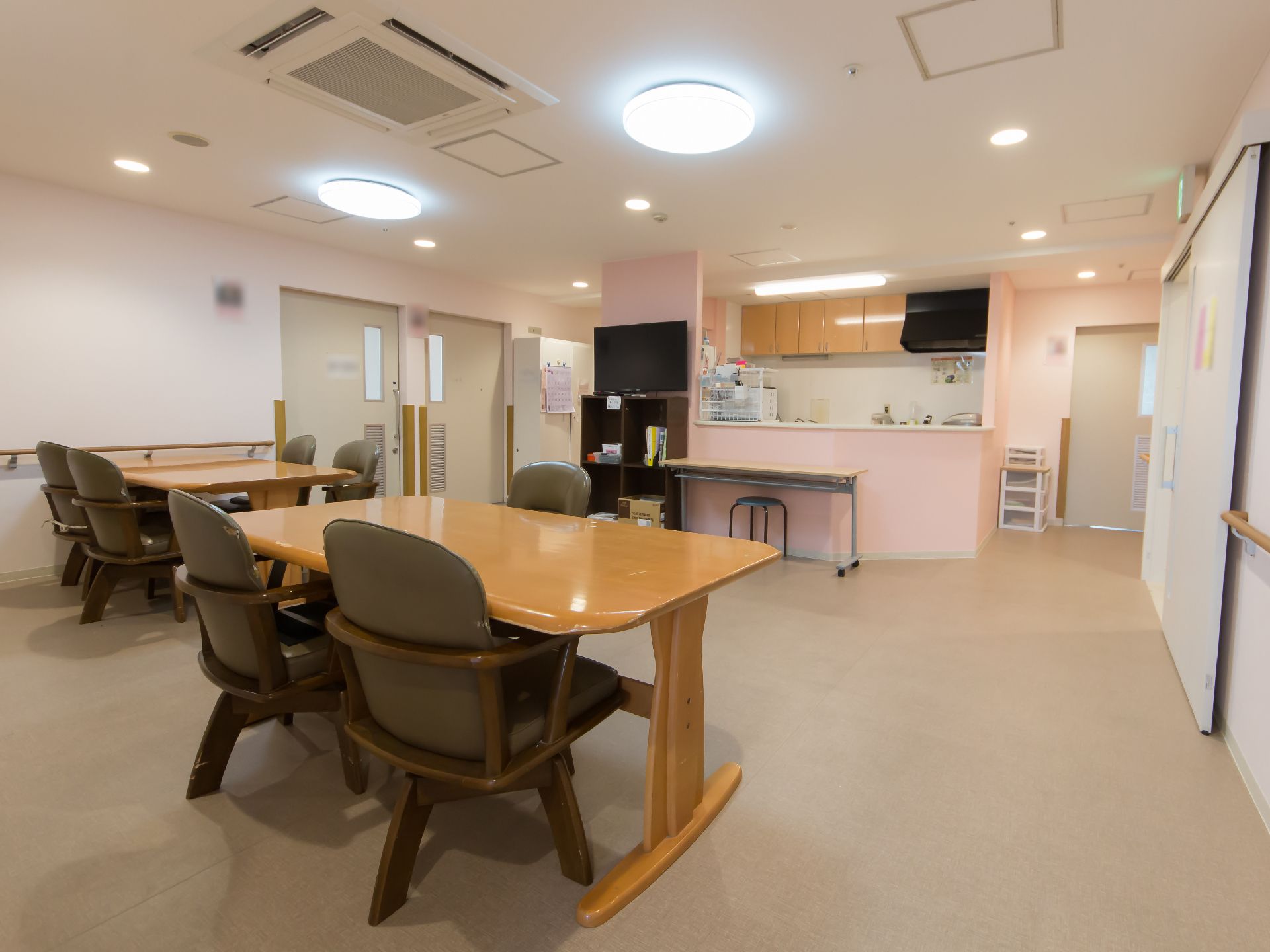 木製のテーブルが２脚と椅子が７脚置かれている。業務用の空調や光度の高い照明が設置されている。奥にはピンク色の壁のキッチンが見える。