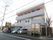 県道に面した３階建ての陸屋根の建物で、３階部分の外壁はオレンジ色である。駐車、駐輪スペースがあり、複数のバイクや車が停まっている。