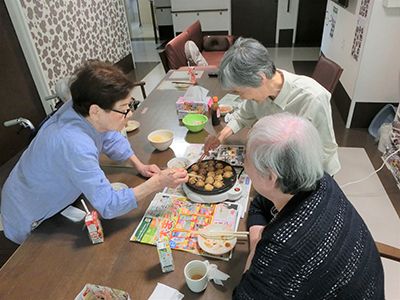 ピンクのカーディガンを着た高齢女性に寄り添い、笑顔で過ごしているピンクのエプロンを付けた若い女性スタッフ。
