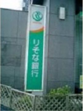 緑の下地に白色で銀行名が書かれた縦長の看板が柱に設置されている。周囲には小さな柵があり、植物が生えている。