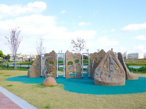 芝生の中に濃い緑色の地面がある公園。広々とした空間があり、大きな岩のオブジェのような遊具などが設置されている。