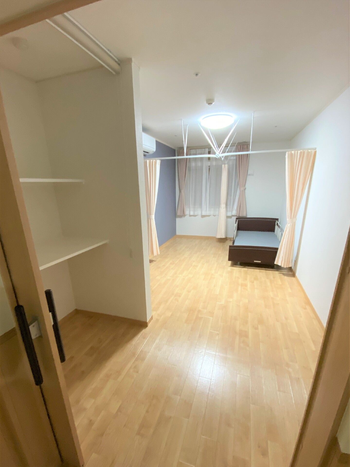 フローリングの部屋には介護ベッドが設置され、仕切り用の薄いオレンジのカーテンも付いている。手前にはオープンタイプの収納棚もある。